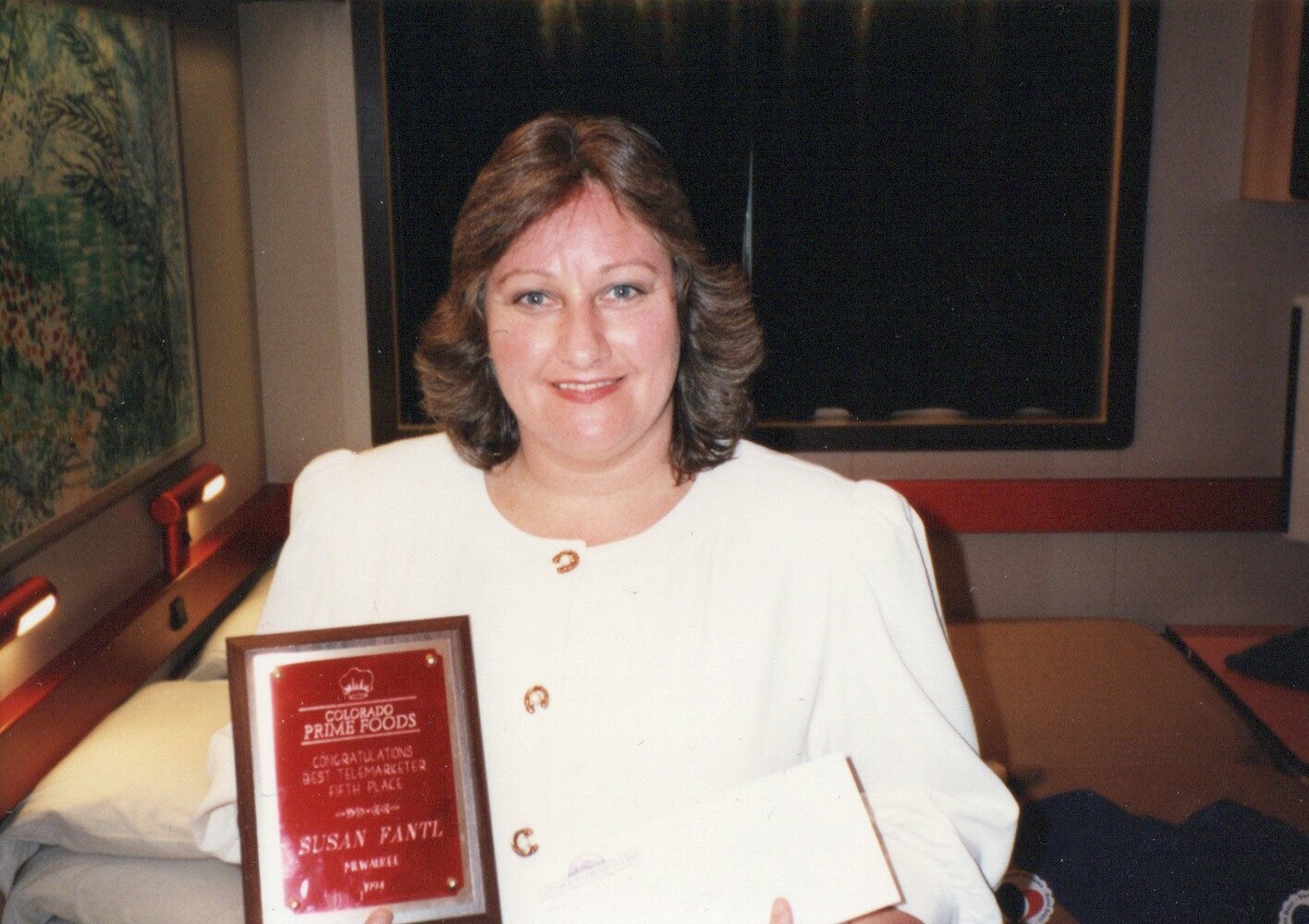 Woman in a white outfit posing with an award plaque and envelope, smiling at the camera in a room.