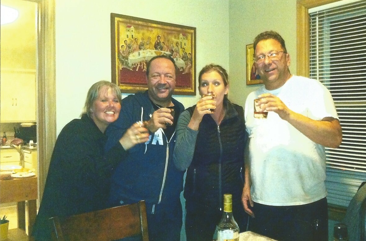 Four adults stand close together, raising glasses in a toast in a cozy dining room, with a bottle on the table in front of them.