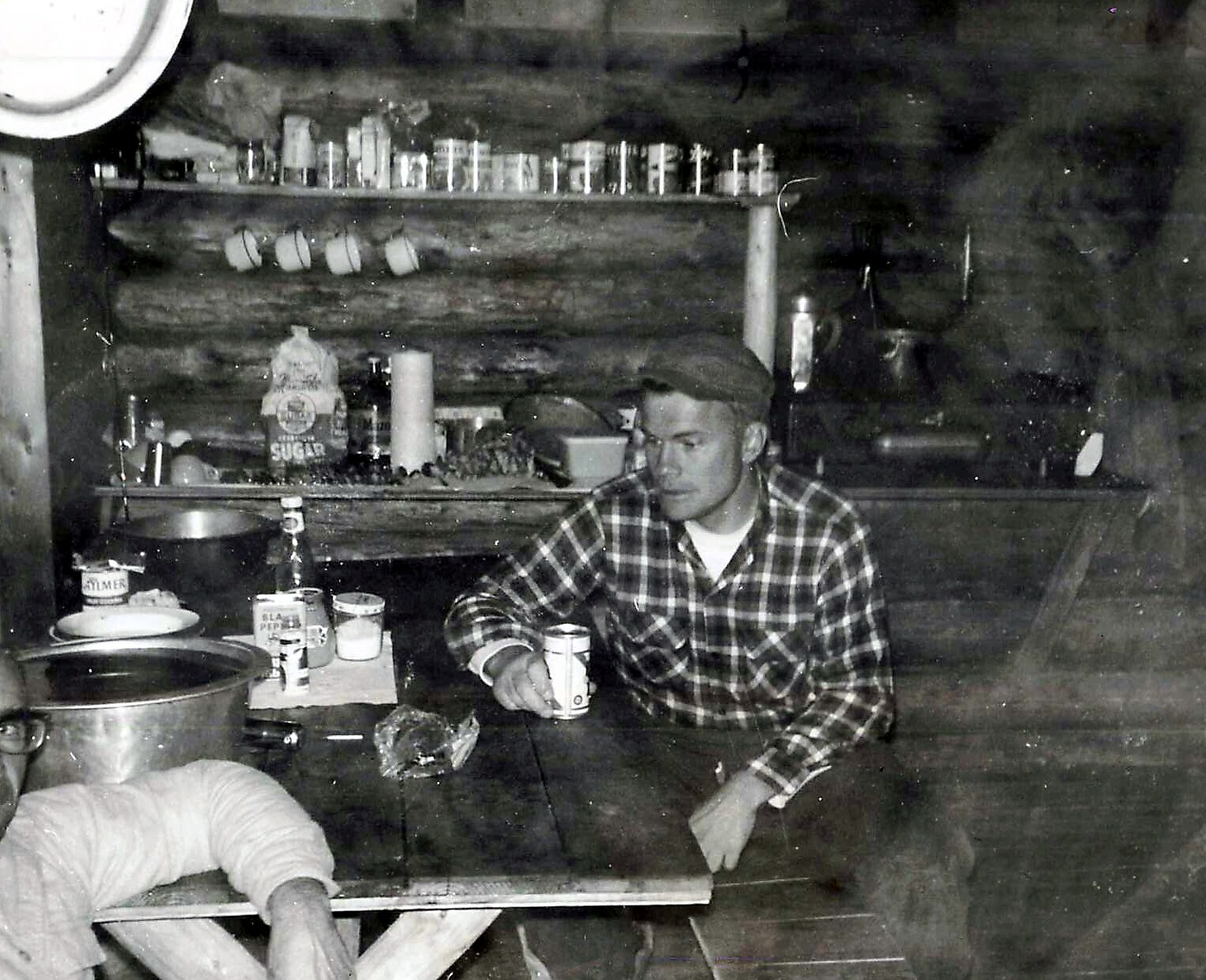 Man in a plaid shirt sits at a rustic cabin table, holding a beer can with shelves of mugs and pantry items behind.