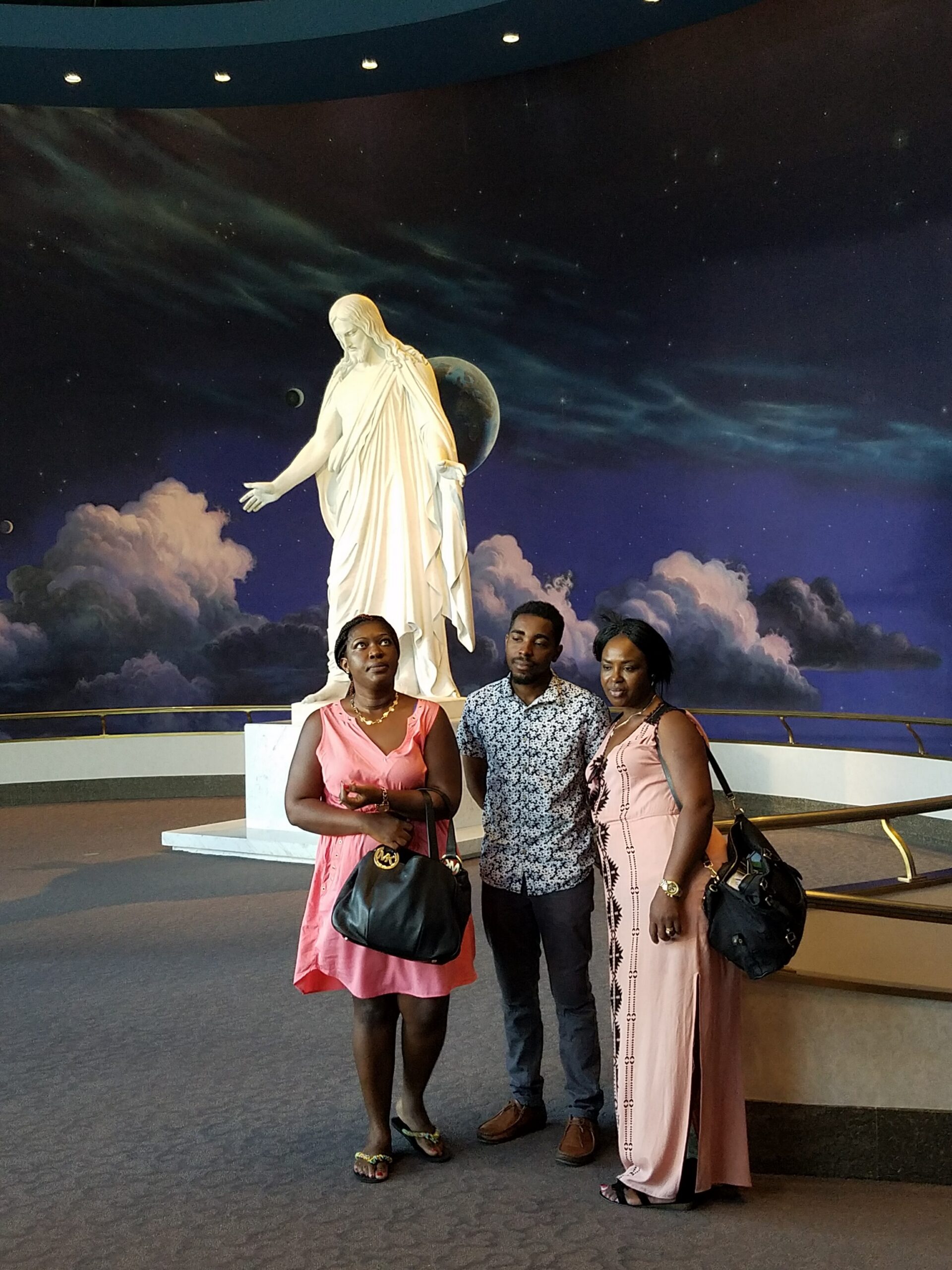 Three adults posing indoors in front of a white statue of Jesus with an outstretched arm, against a starry, cloudy backdrop.