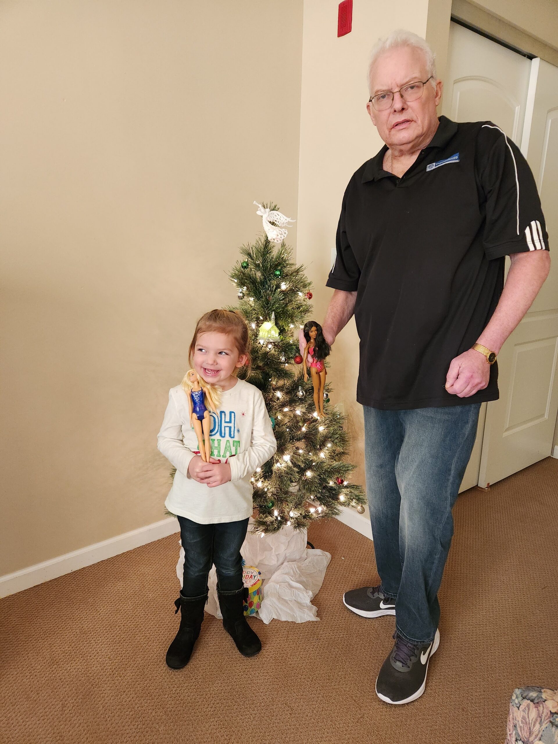 Young girl smiling while holding two Barbie dolls next to a small decorated Christmas tree indoors with a man nearby.