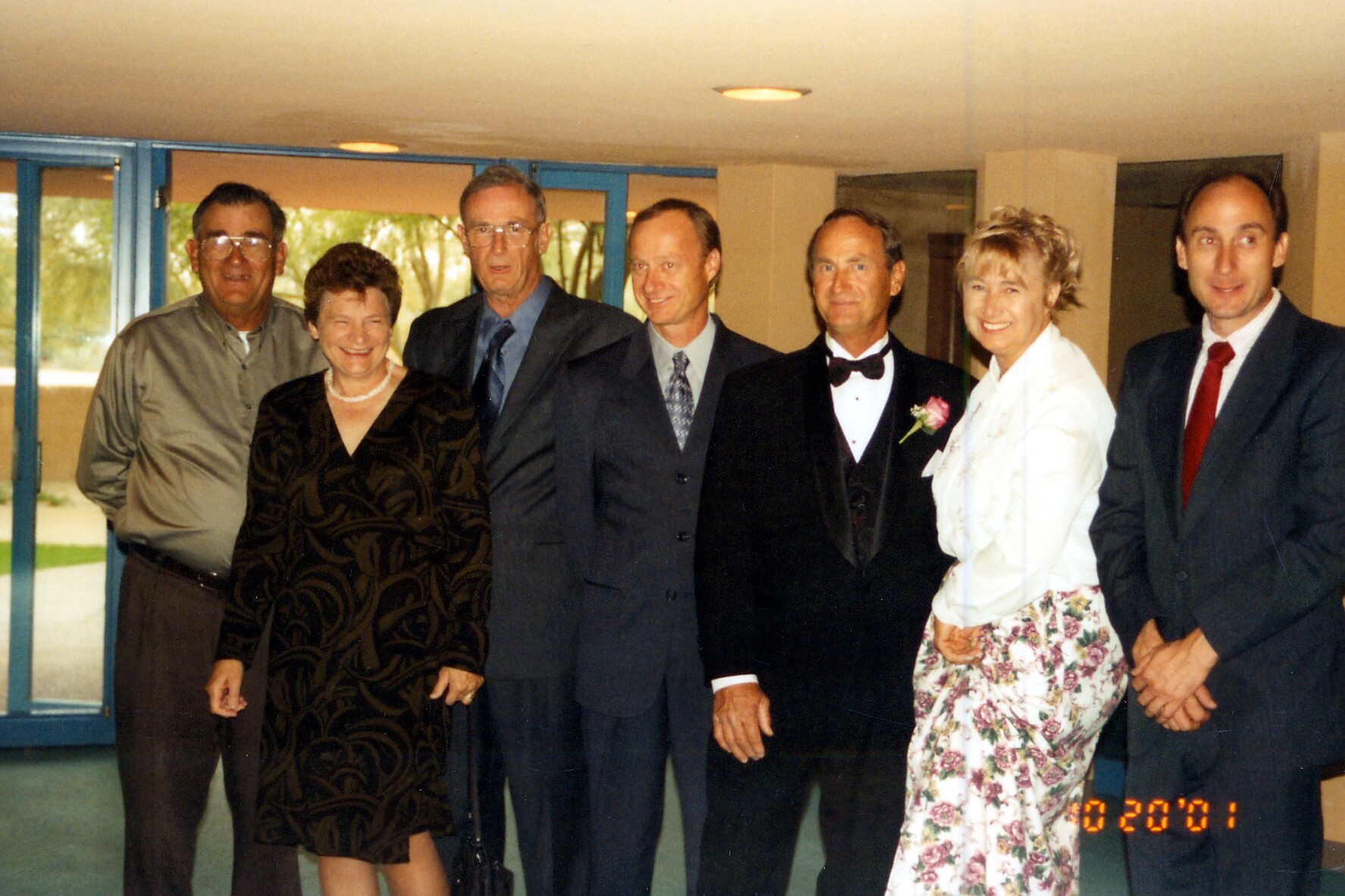 Seven adults in formal attire pose together indoors for a group photo at a social event, smiling.