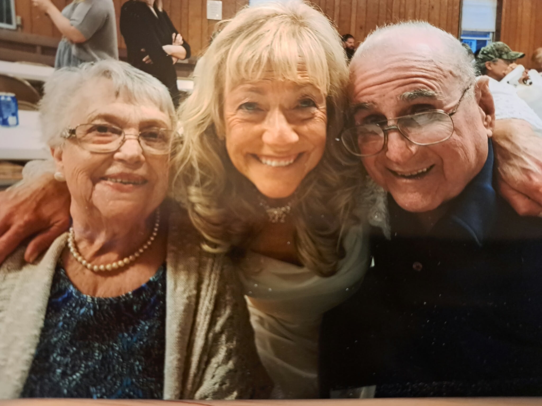 Three seniors smiling for a selfie indoors at a gathering: left woman with glasses and pearl necklace, center woman, right man with glasses.