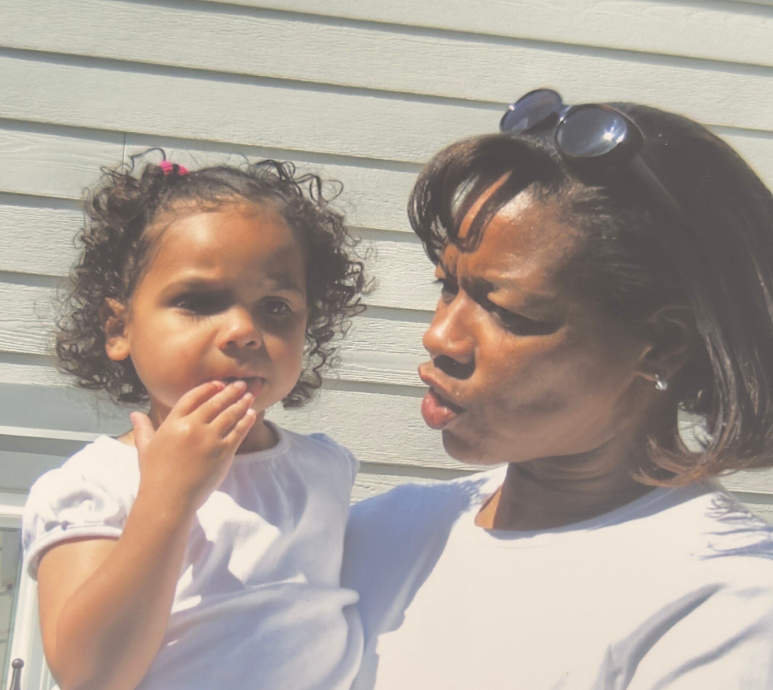 Close-up of a young child with curly hair and a smiling adult wearing sunglasses on a sunny day outdoors.