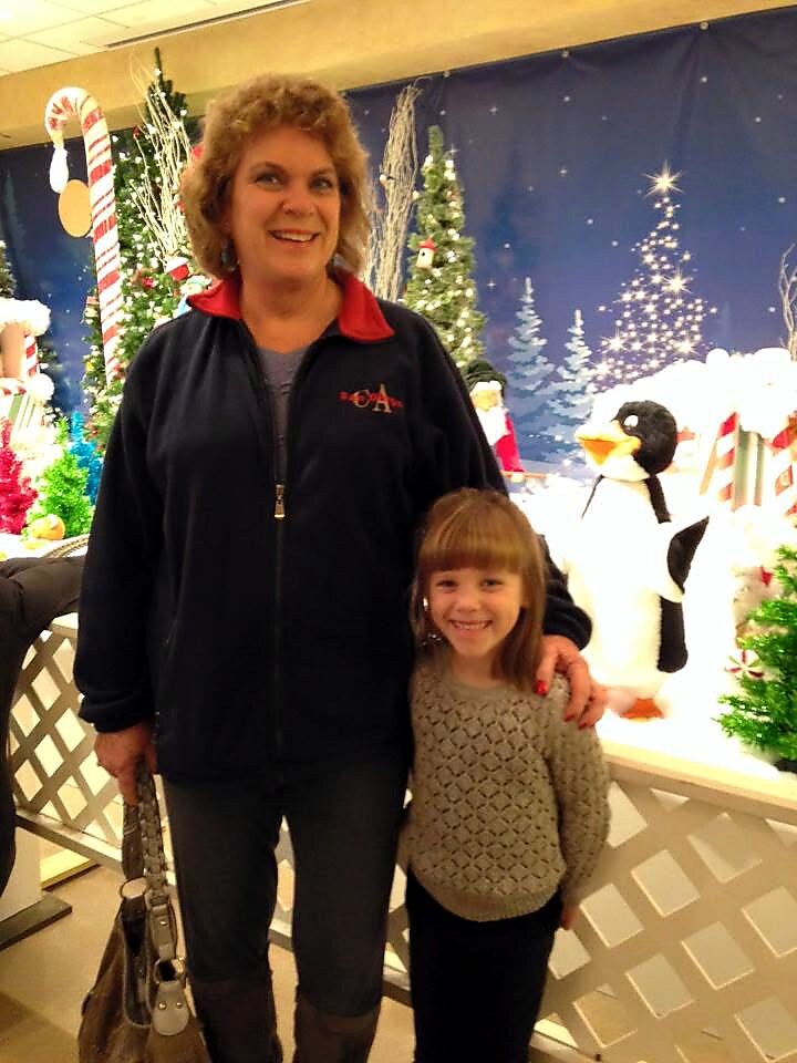 Woman with short curly hair stands beside a smiling young girl in front of a festive Christmas display with candy canes, trees, and a penguin figure in the background.