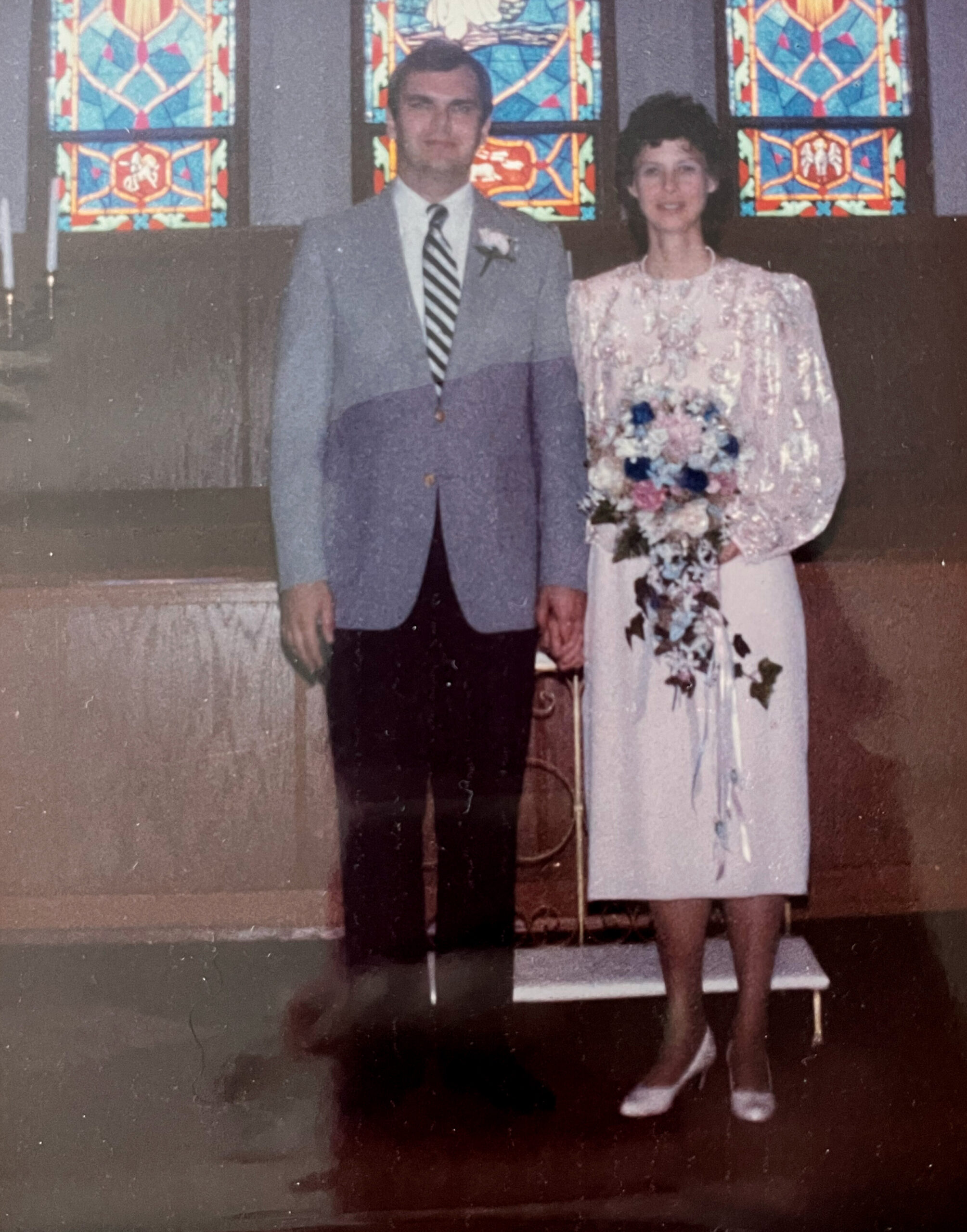 Bride and groom stand together in front of colorful stained glass at a church; man in a light gray suit with striped tie and boutonniere, woman in a pale pink floral dress holding a bouquet.