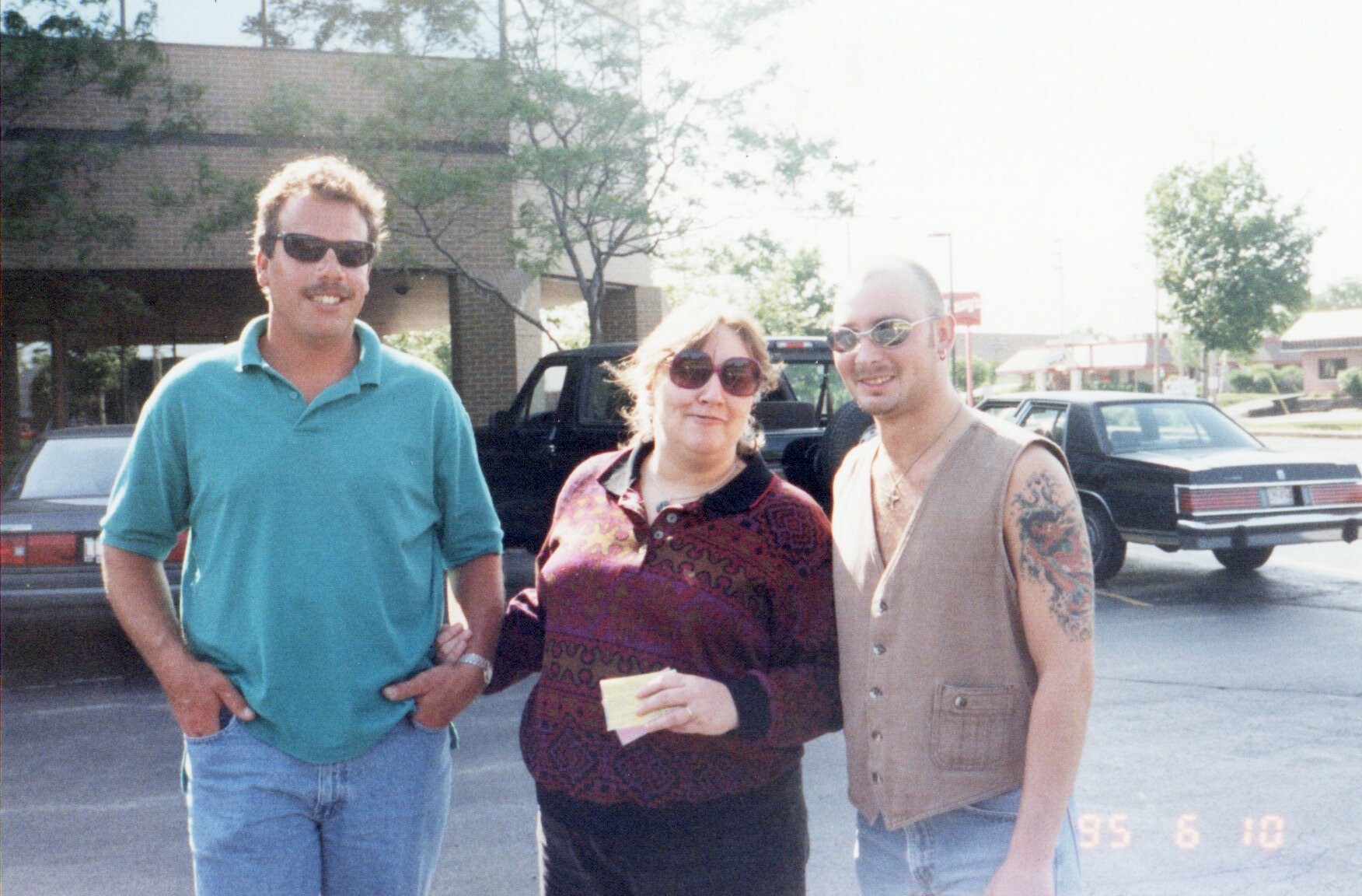 Three adults posing in a parking lot: man in turquoise polo, woman in burgundy patterned sweater, and man in beige sleeveless vest with sunglasses; a black pickup truck behind them.