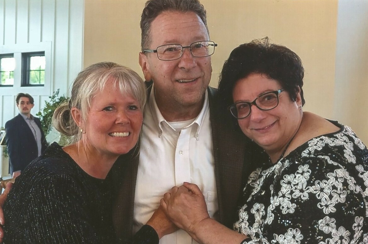 Three adults posing together and smiling, two women on either side of a man in a suit, arms around each other at an indoor event.