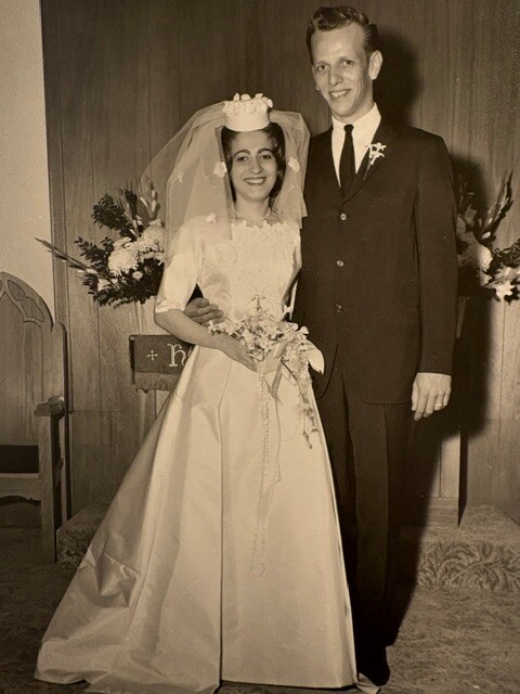 Bride in a white wedding gown and veil holds a bouquet, standing with a man in a dark suit under church decor, a vintage wedding photo.