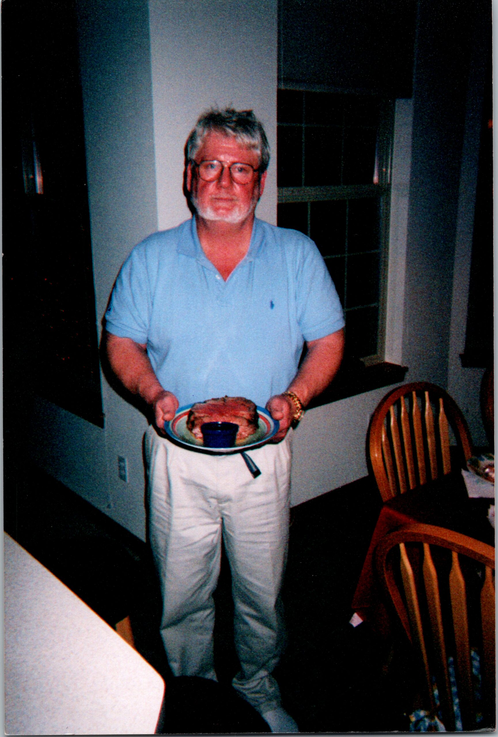 Older man with gray hair and beard in a light blue polo holds a plate with a sandwich and a blue cup at a dining table.