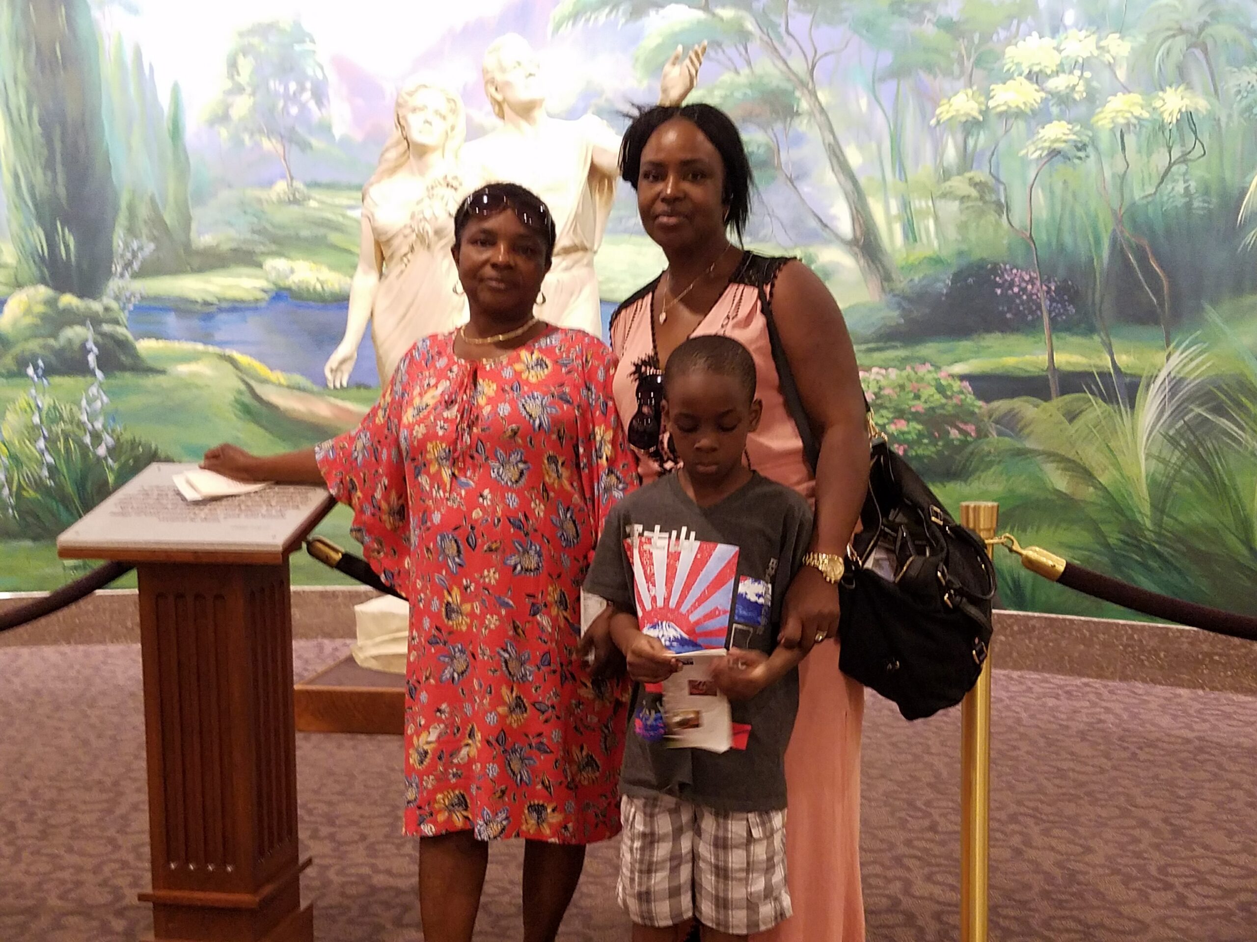 Three people posing together in front of a large nature mural; a woman in a red floral dress holds a pamphlet while a woman in pink stands beside a young boy holding papers.