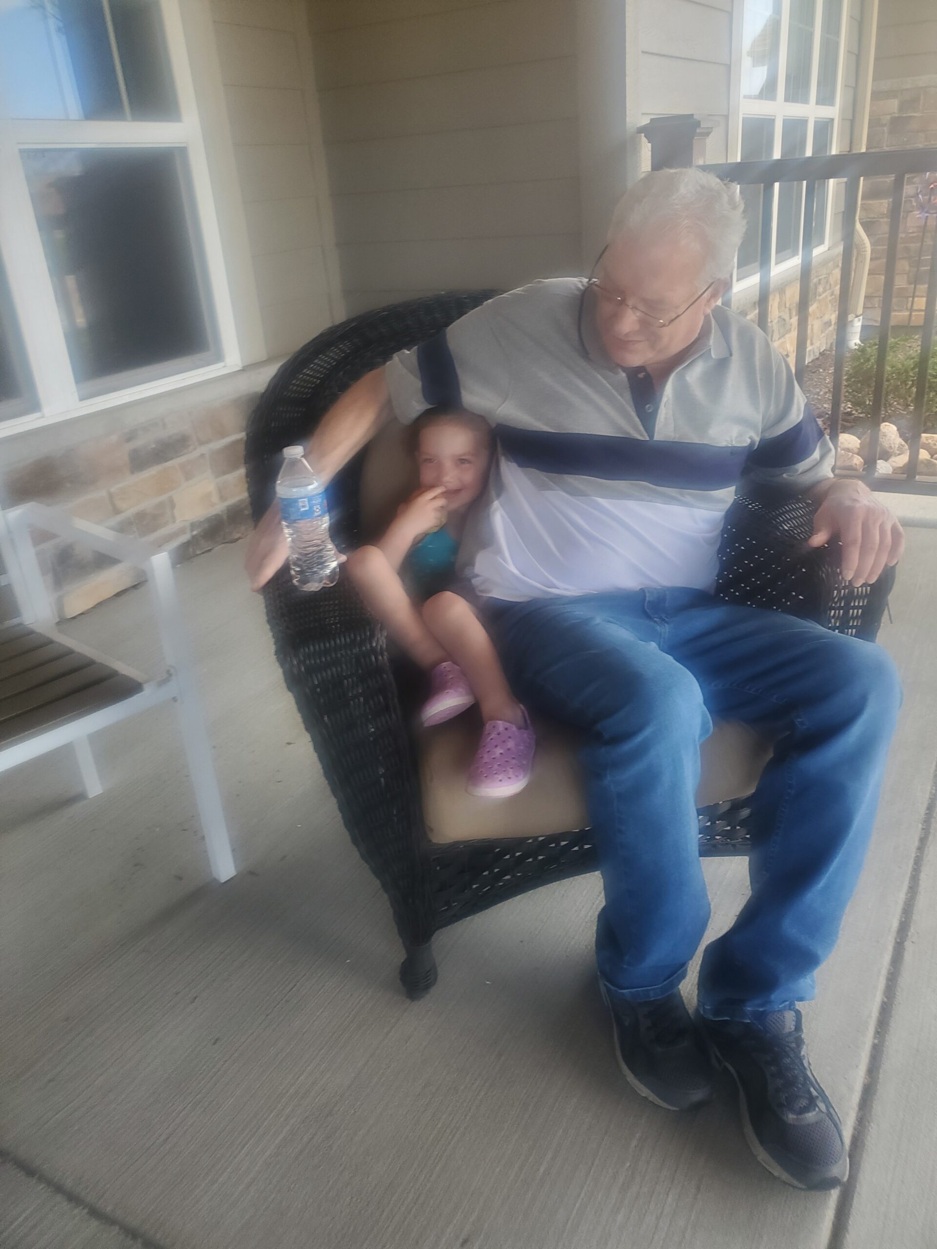 Older man with white hair and glasses sits in a wicker chair on a porch, a young girl on his lap holding a water bottle nearby.