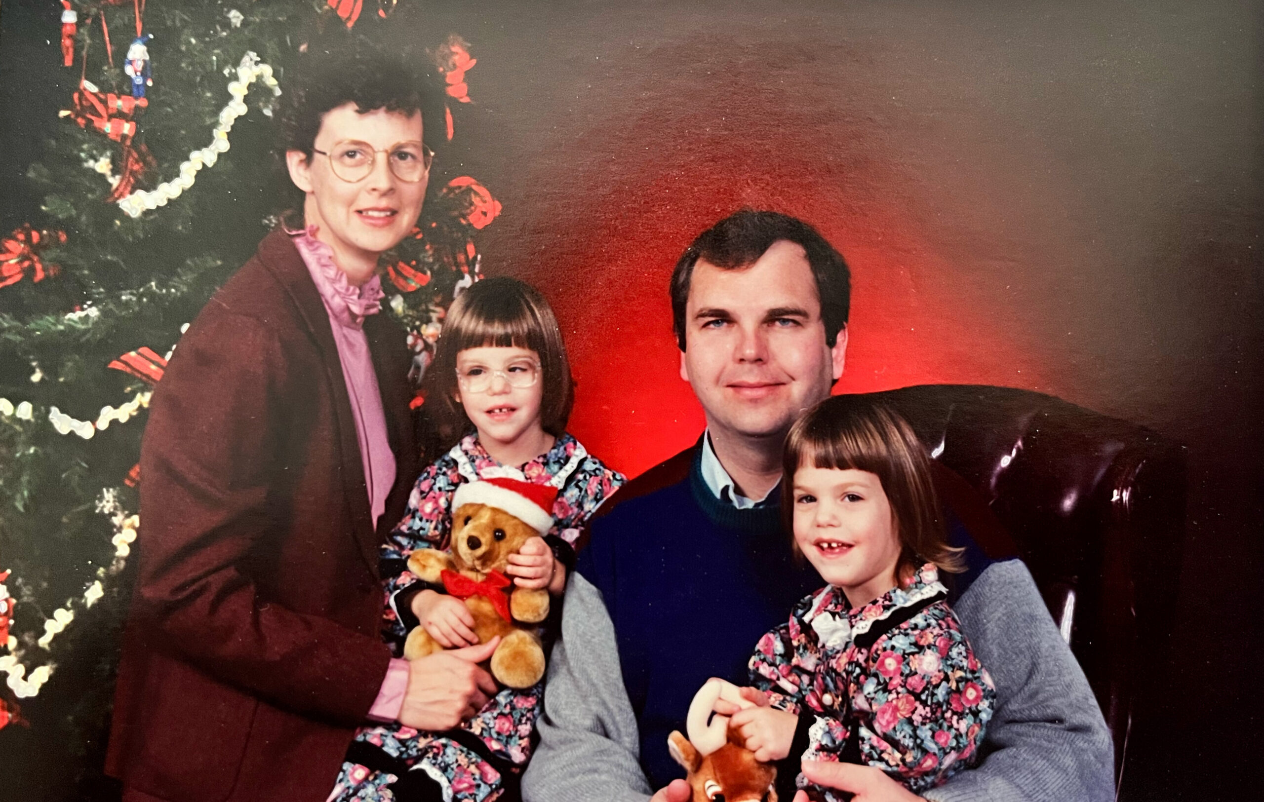 Family portrait by a Christmas tree: mother, father, and three children posing with plush bears and festive outfits.
