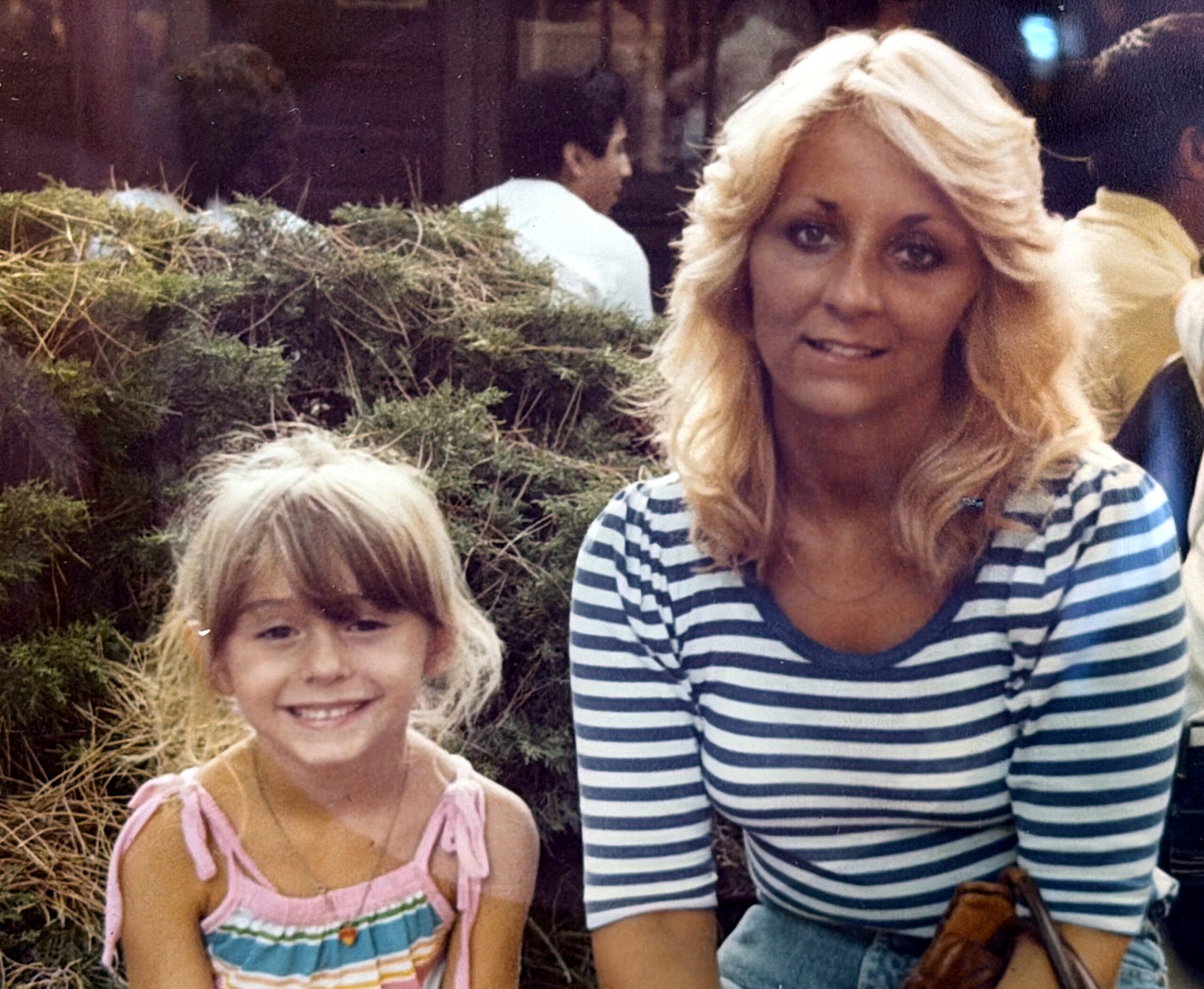 Two people posing outdoors: a smiling woman in a blue-and-white striped shirt next to a smiling young girl in a pink dress, with evergreen bushes in the background.