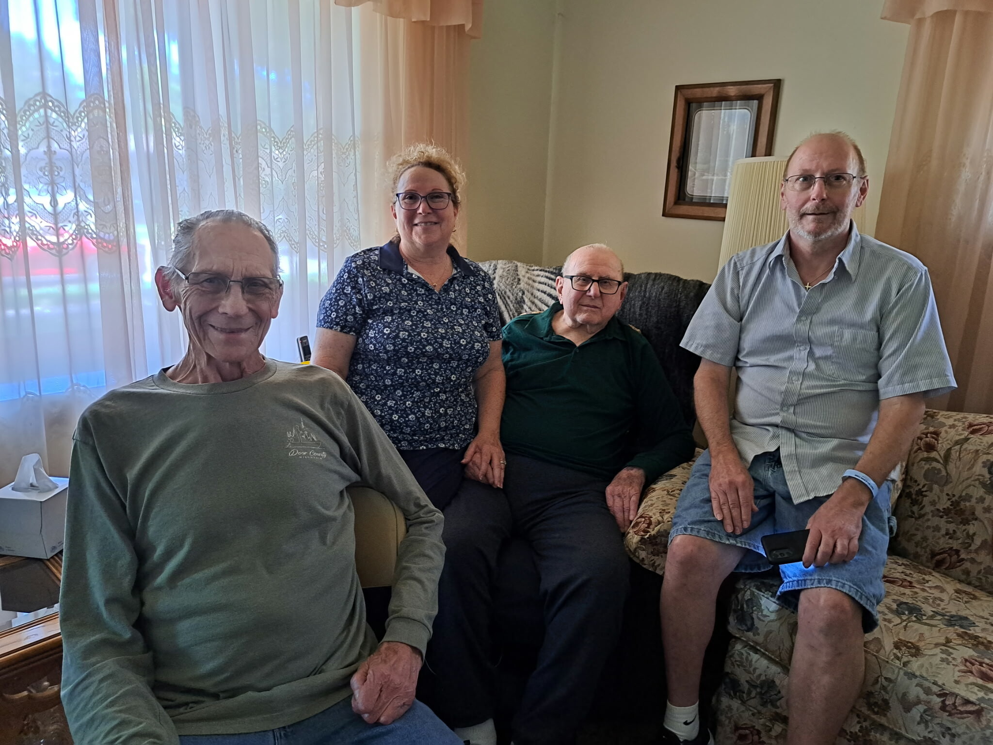 Four adults seated on a floral sofa in a cozy living room, smiling at the camera by a lace-curtained window.