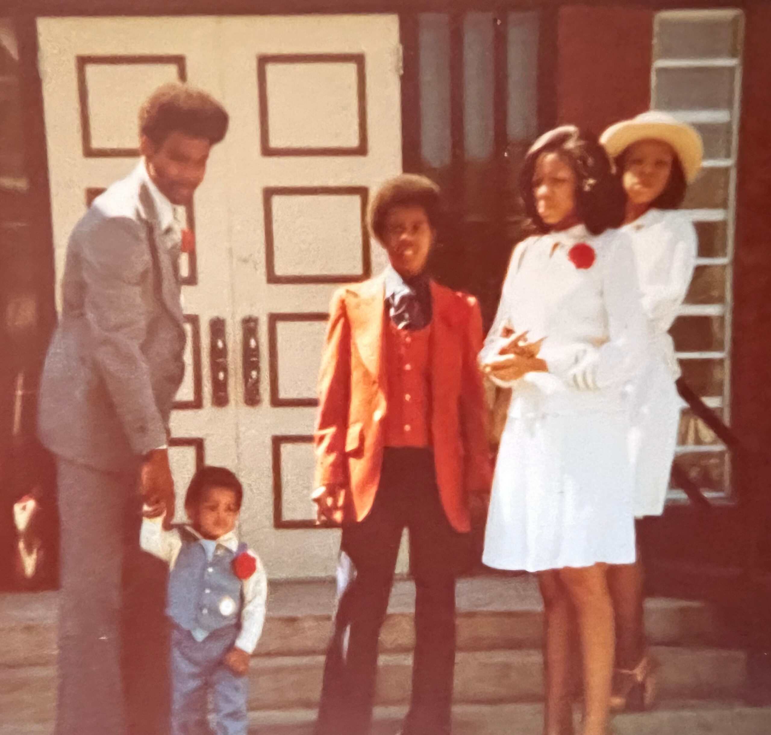 A small group of people posing outdoors in front of a house, wearing 1970s-style clothing.