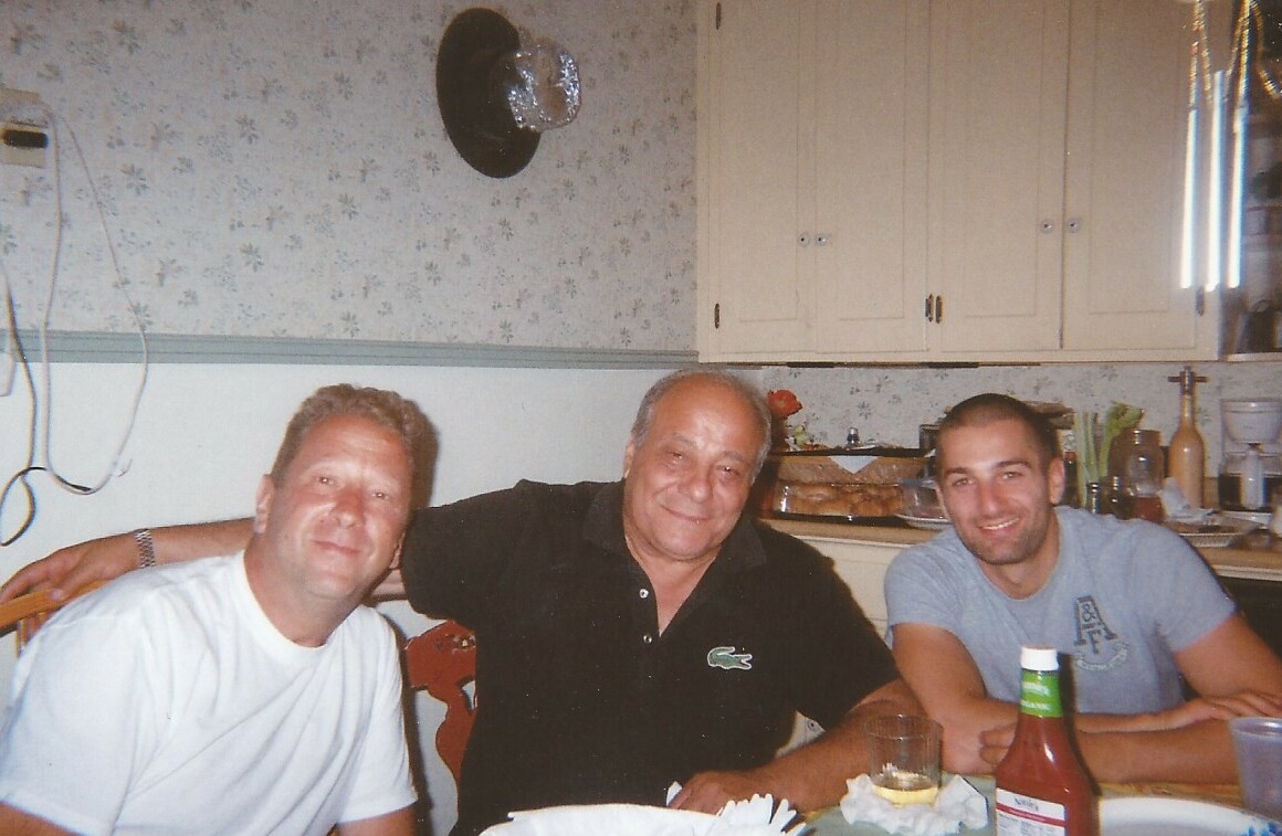 Three men sit at a kitchen table smiling for the camera, with drinks and condiments on the table in front of them.