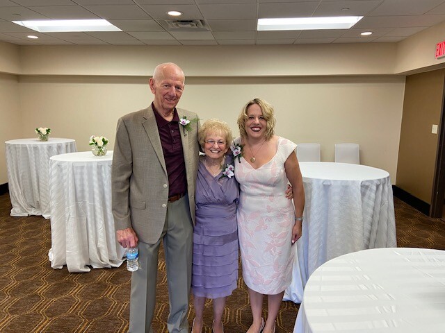 Three people posing at a reception: man in a gray suit, an elderly woman in a lavender dress, and a woman in a light pink dress; corsages on their outfits with white round tables in the background.
