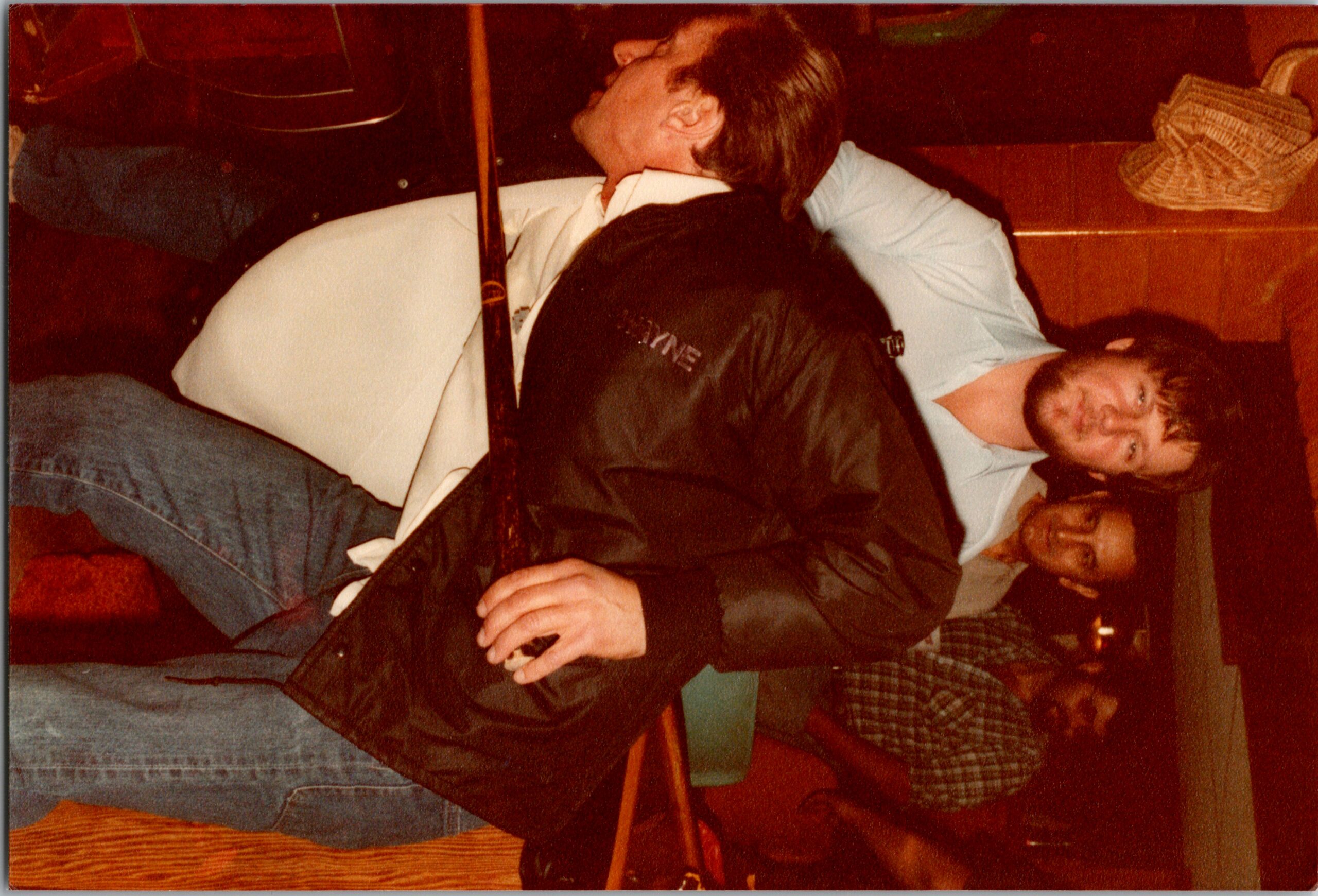 Group of men hanging out indoors, some leaning on a wooden railing and smiling at the camera.