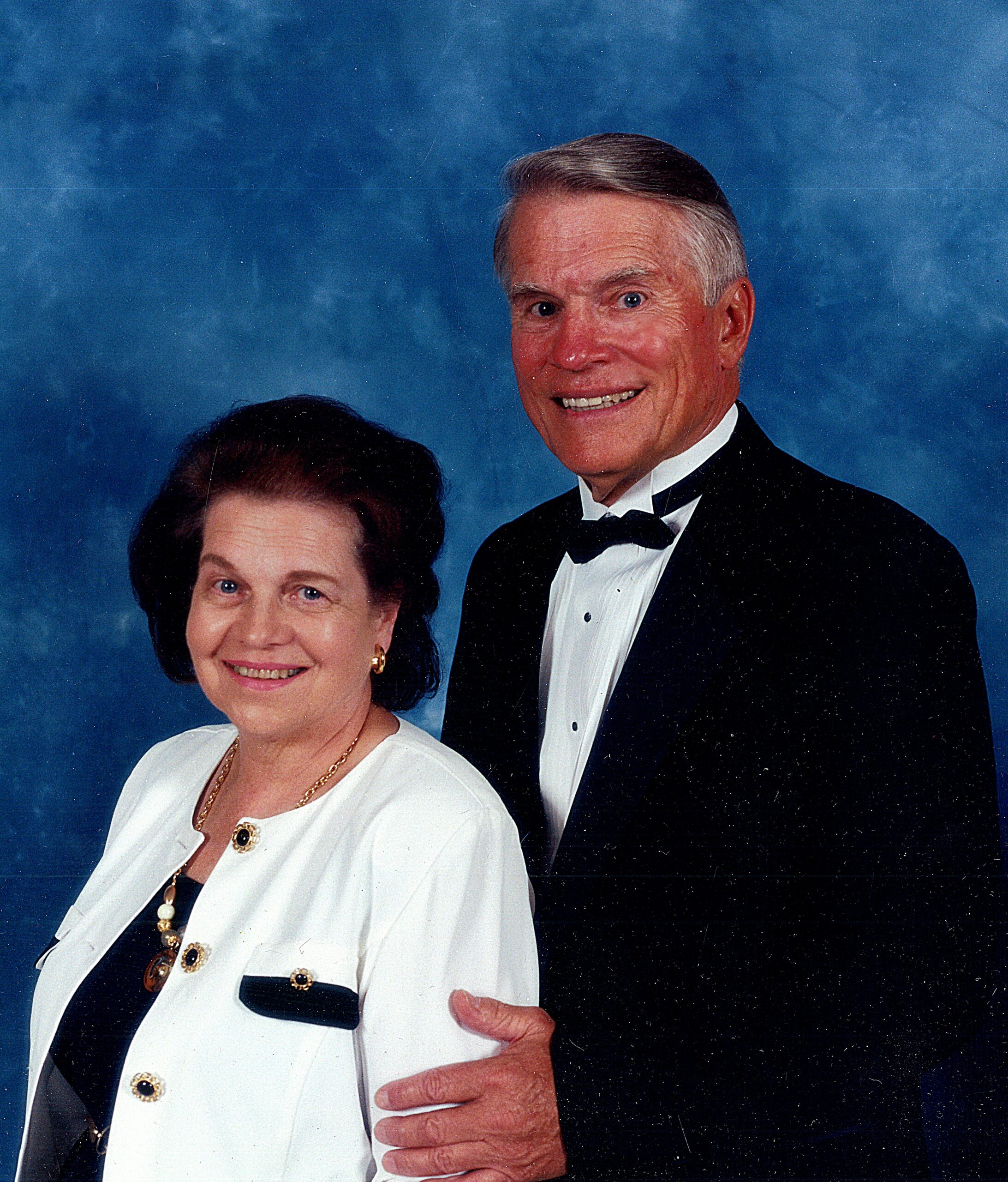 Smiling senior couple in formal attire posing against a blue studio backdrop.