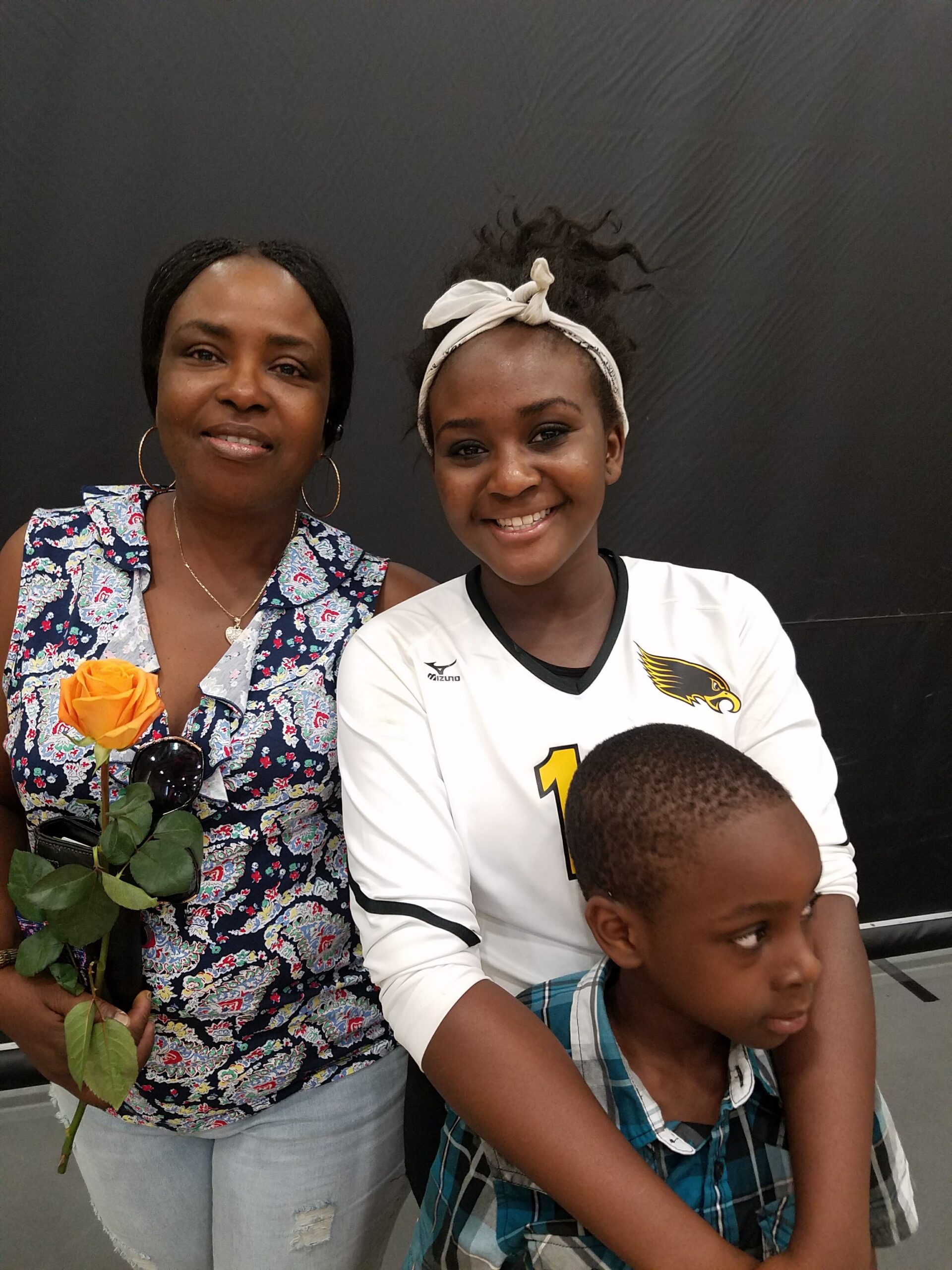 Two women posing together: an older woman in a floral shirt holds a yellow rose and sunglasses, smiling at the camera against a dark backdrop.