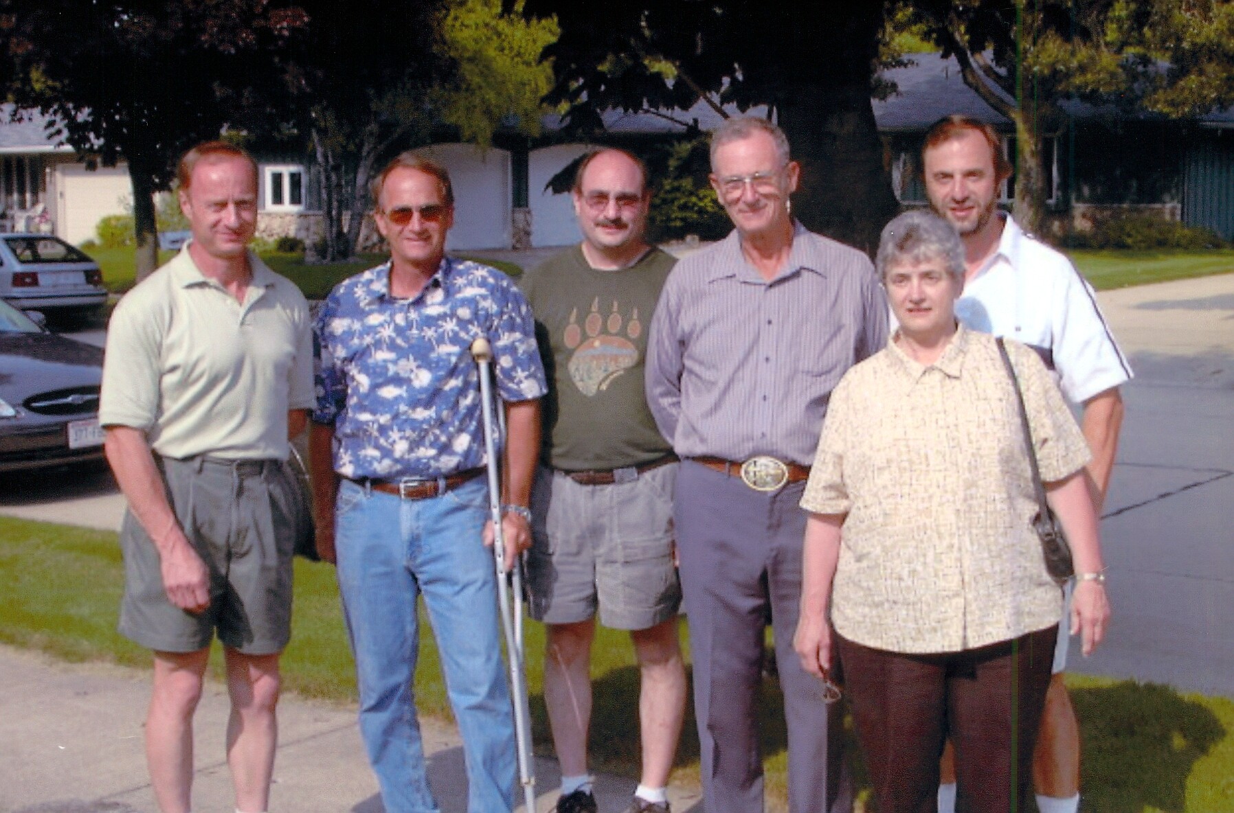 Six adults pose for a casual group photo on a suburban street, with a woman in front and five men behind them, sunny day, casual attire. One man uses crutches for support beside purple shorts and a patterned shirt.