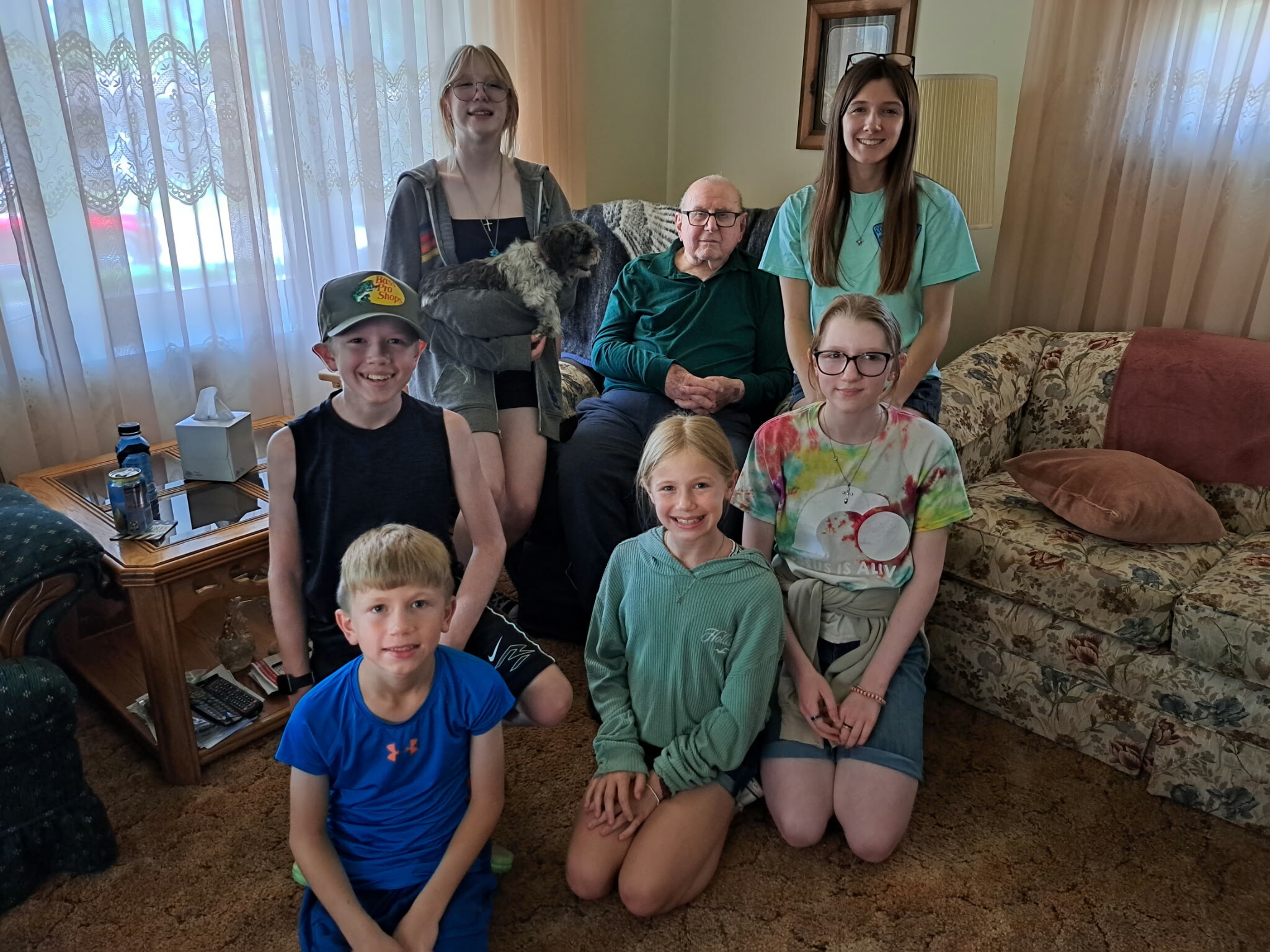 Multi-generational family photo in a living room: elderly man on the sofa with kids gathered around and two young women standing behind; a small dog sits on a girl’s lap at back.