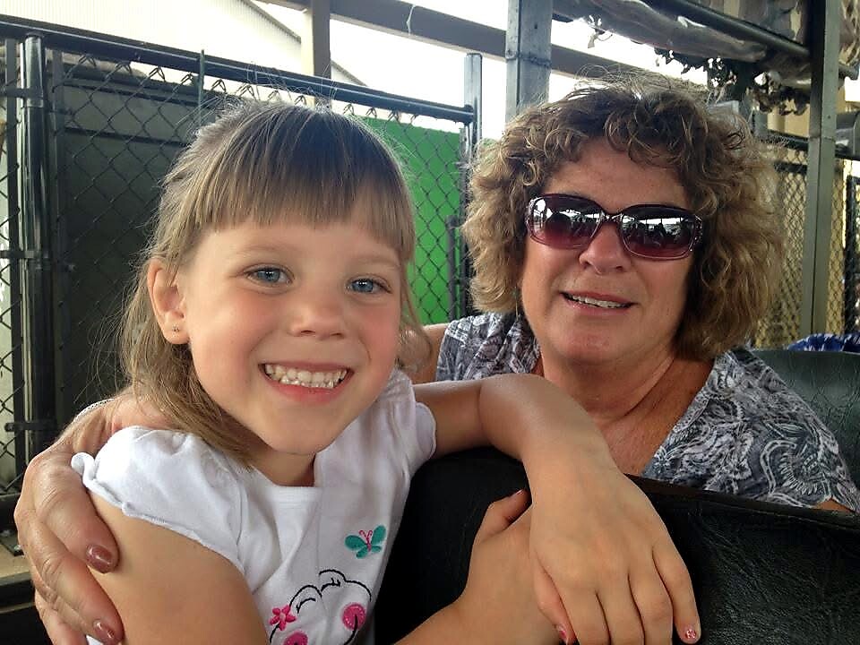 Smiling young girl with bangs hugs an older woman in sunglasses on a ride, with a chain-link fence in the background.|Moments shared between granddaughter and grandmother during a sunny outing.
