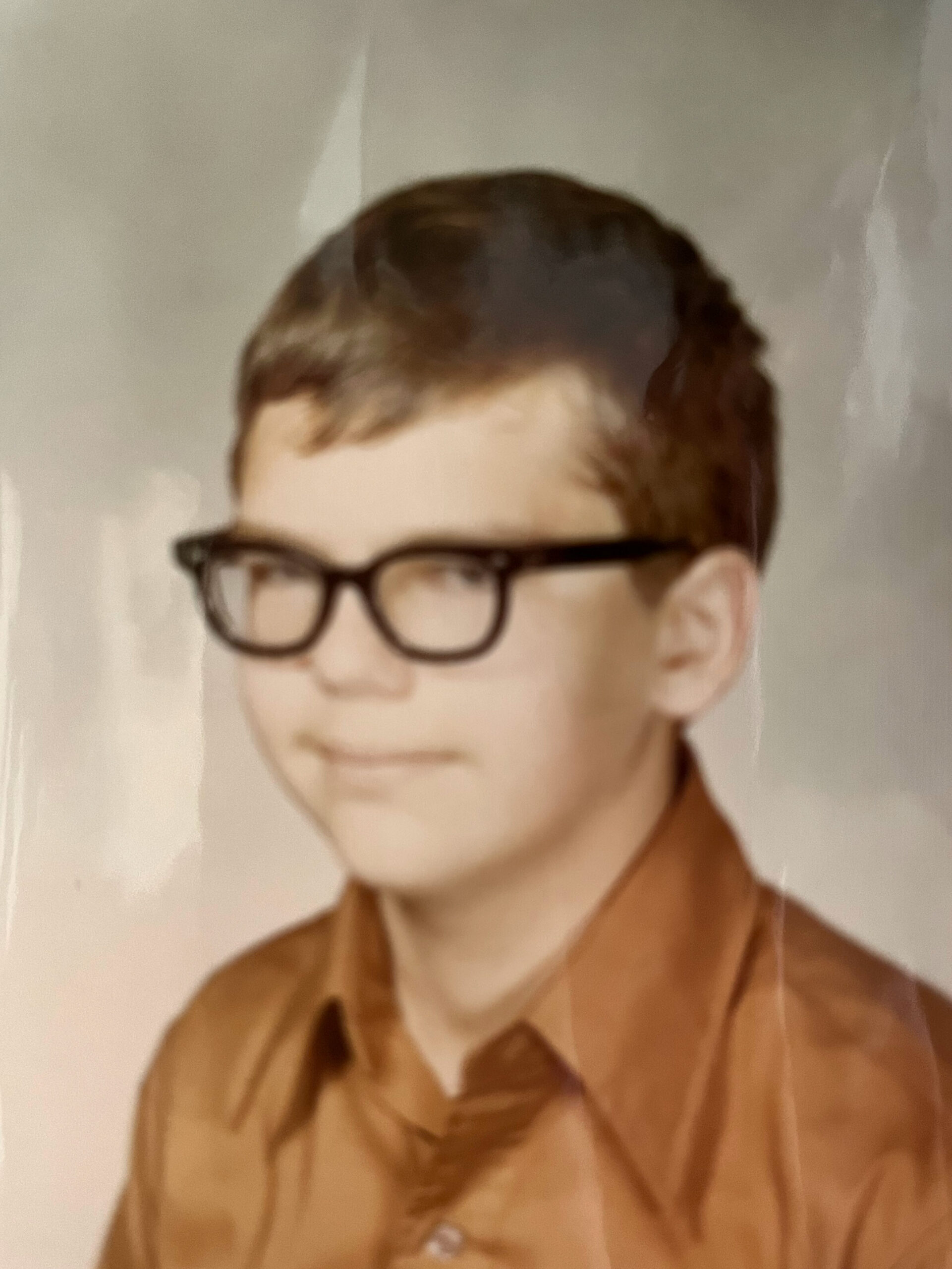 Close-up portrait of a person with short brown hair and dark glasses wearing a brown shirt, against a neutral background.