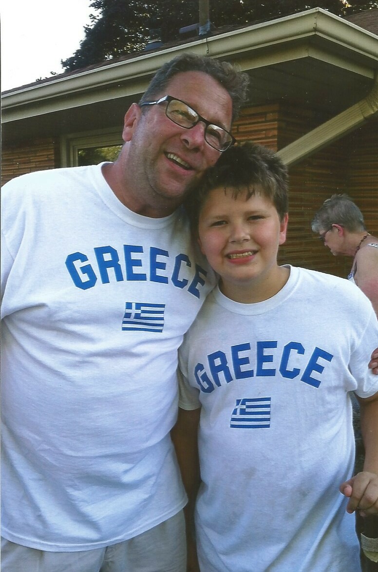 Two people, a man and a boy, wearing matching white Greece T-shirts with a small Greek flag, smiling outdoors beside a house in the background.
