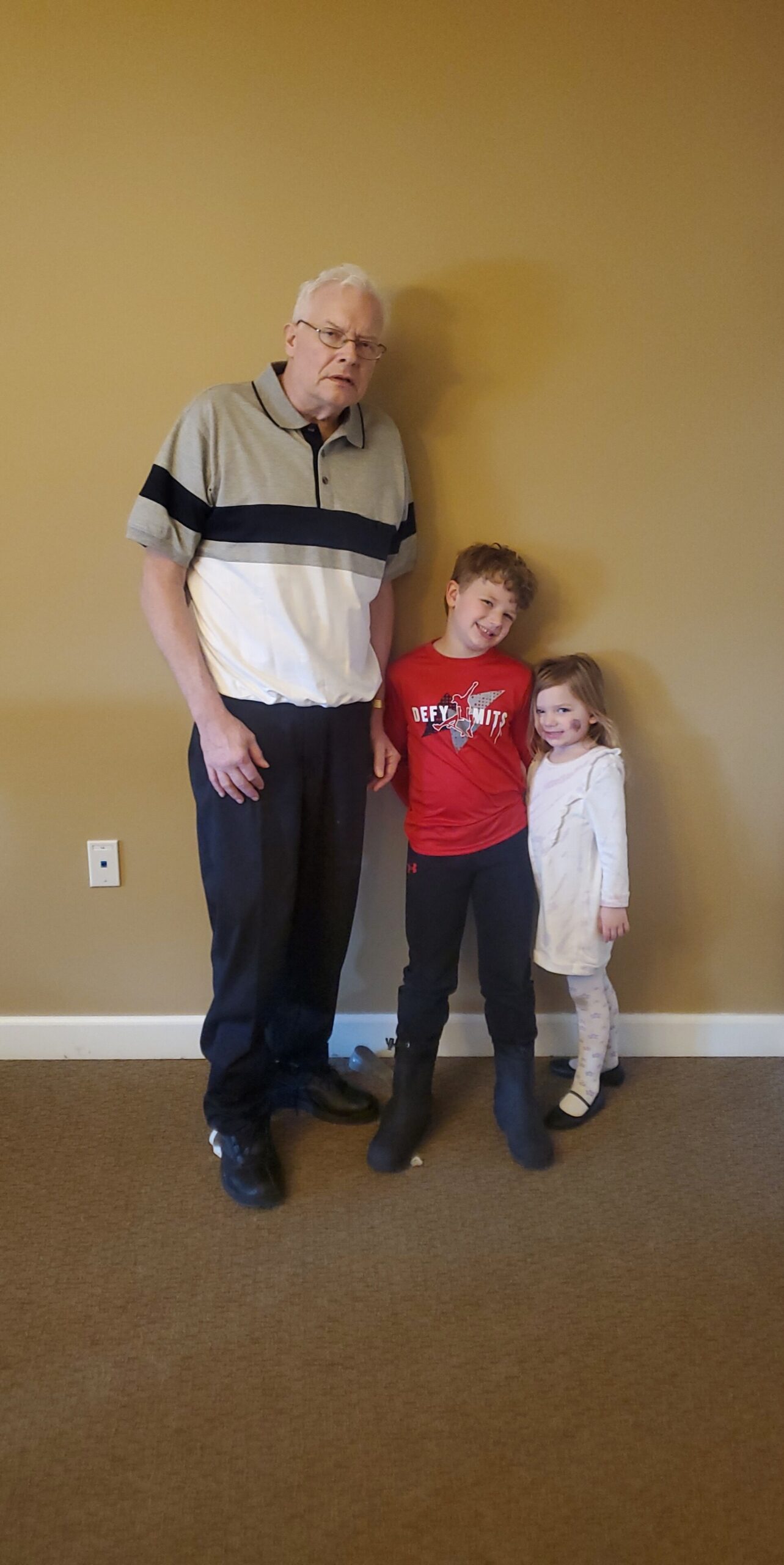Older man with white hair and glasses stands with a smiling boy in a red shirt and a girl in a white dress, posing against a beige wall indoors.