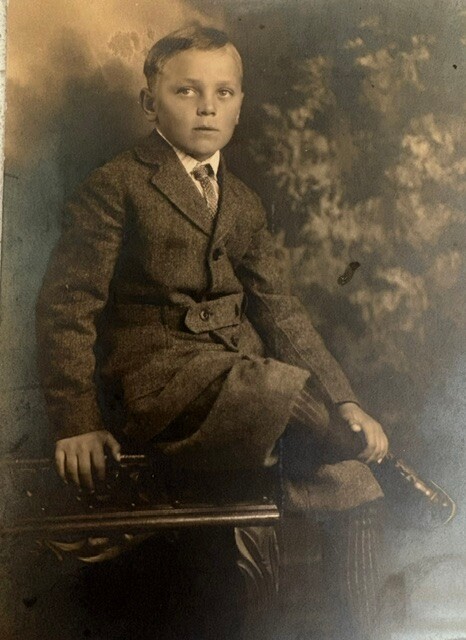 Young boy in a formal suit seated on a wooden bench in a studio portrait, with a neutral backdrop behind him.
