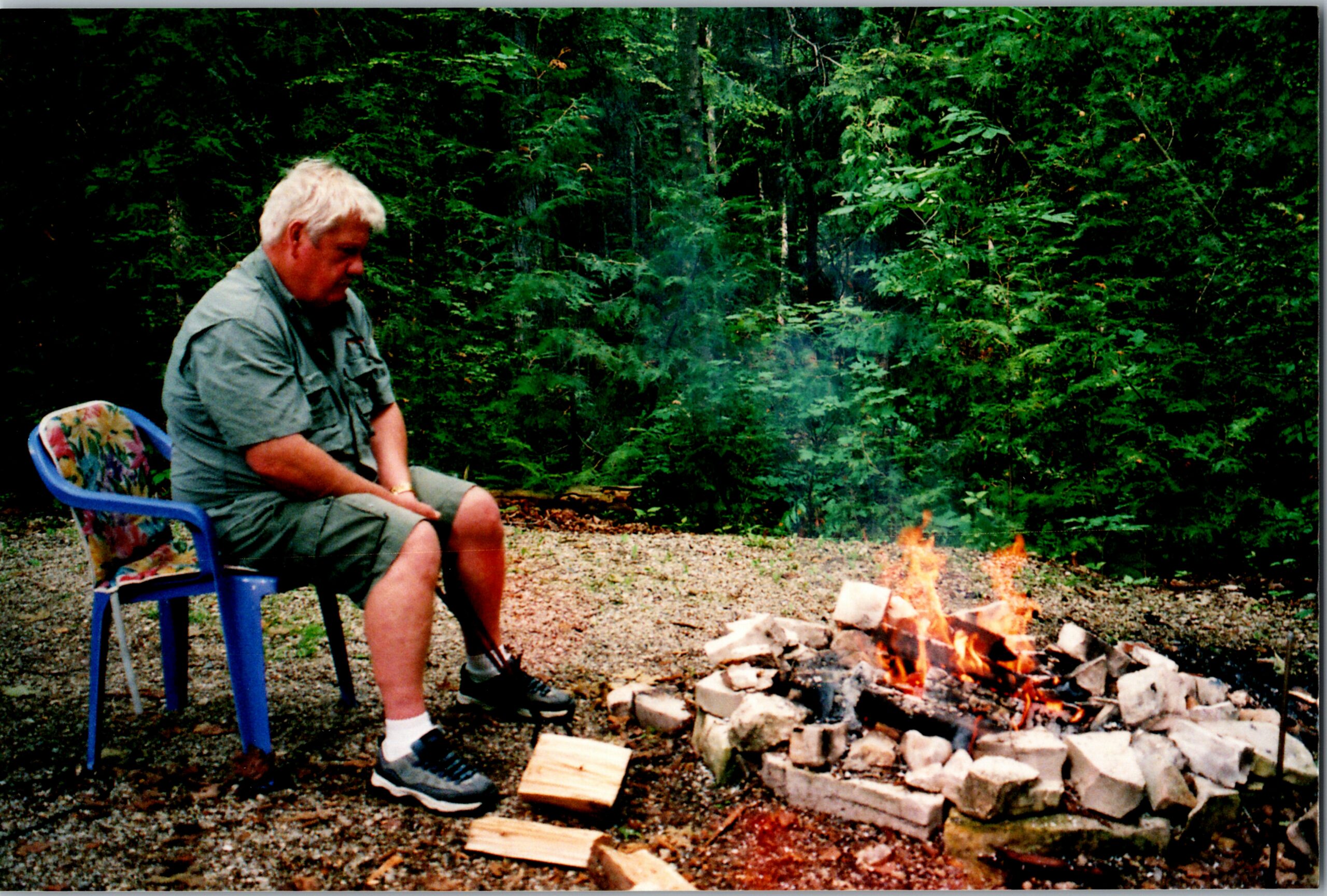Older man sitting on a blue chair by a campfire in a wooded area, with bricks arranged around the flames and wood on the ground nearby