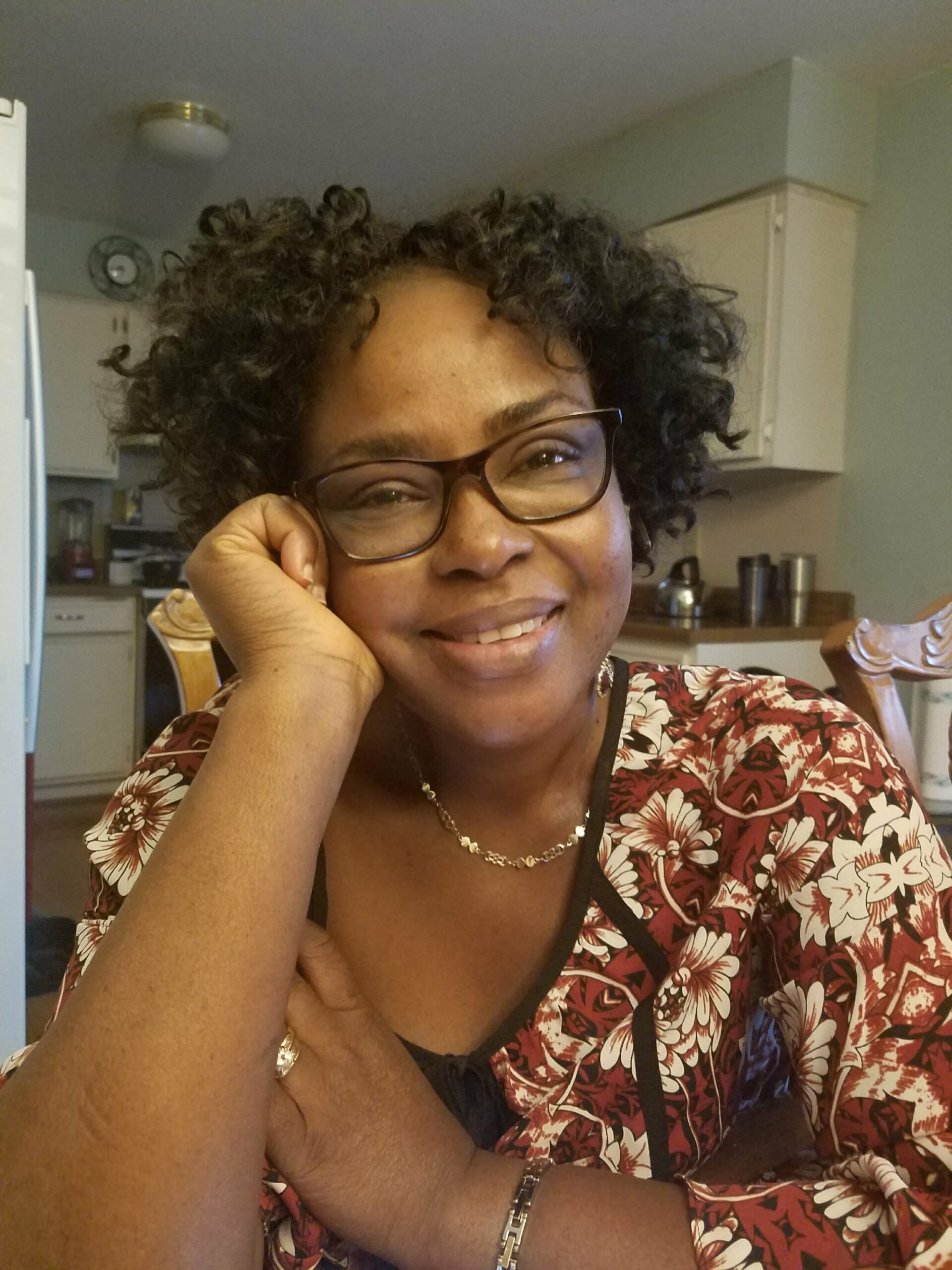 Smiling woman with glasses rests her chin on her hand in a home kitchen, wearing a red floral blouse and a necklace.