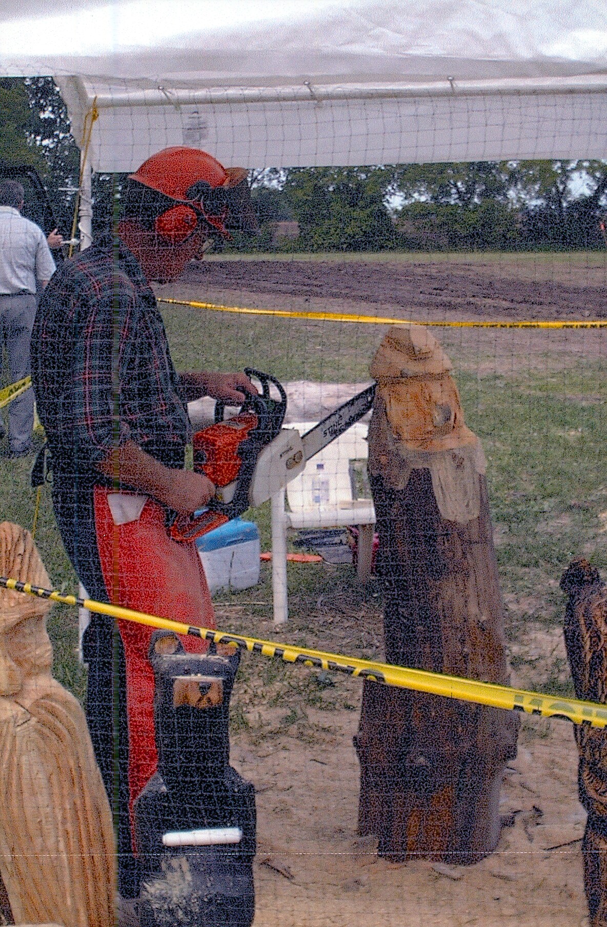 Man in an orange helmet and plaid shirt operating a chainsaw to cut a log under a white canopy, with yellow safety tape in the foreground.