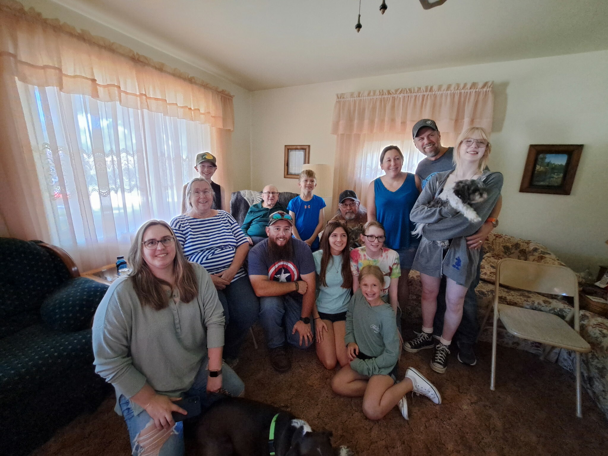 Group photo of a large family and friends indoors in a living room, with a black dog in the foreground and pink curtains in the background.