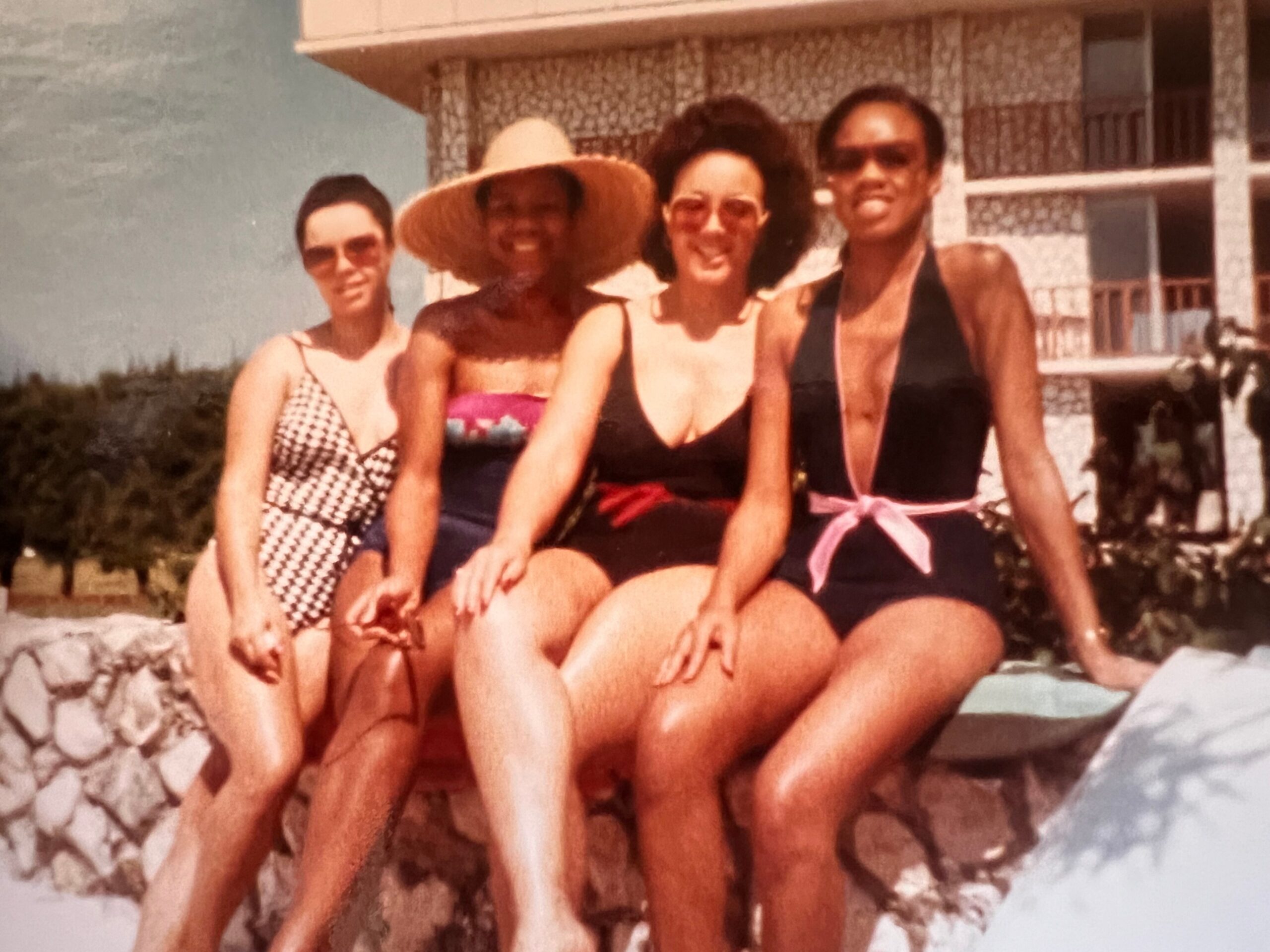 Four women in swimsuits posing together outdoors, with a building and rocky foreground in the background.