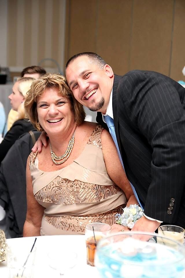 Smiling couple at a formal event, man in a suit leaning over a seated woman in a beige dress with a beaded necklace.