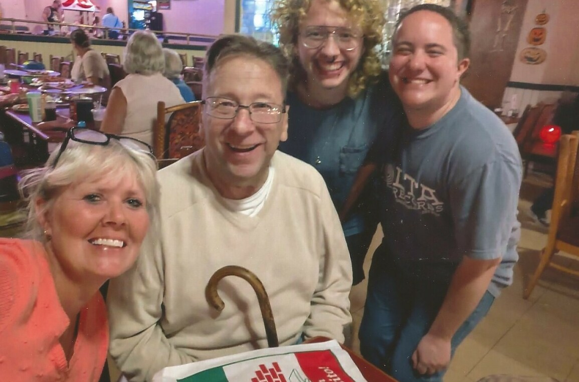 Four friends posing for a selfie in a restaurant; center man with a cane smiles as others lean in and smile behind him.