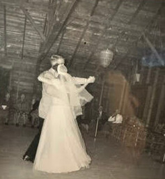 Vintage photo of a bride in a white gown dancing with a man in a suit in a large indoor hall, giving a celebratory wedding vibe.