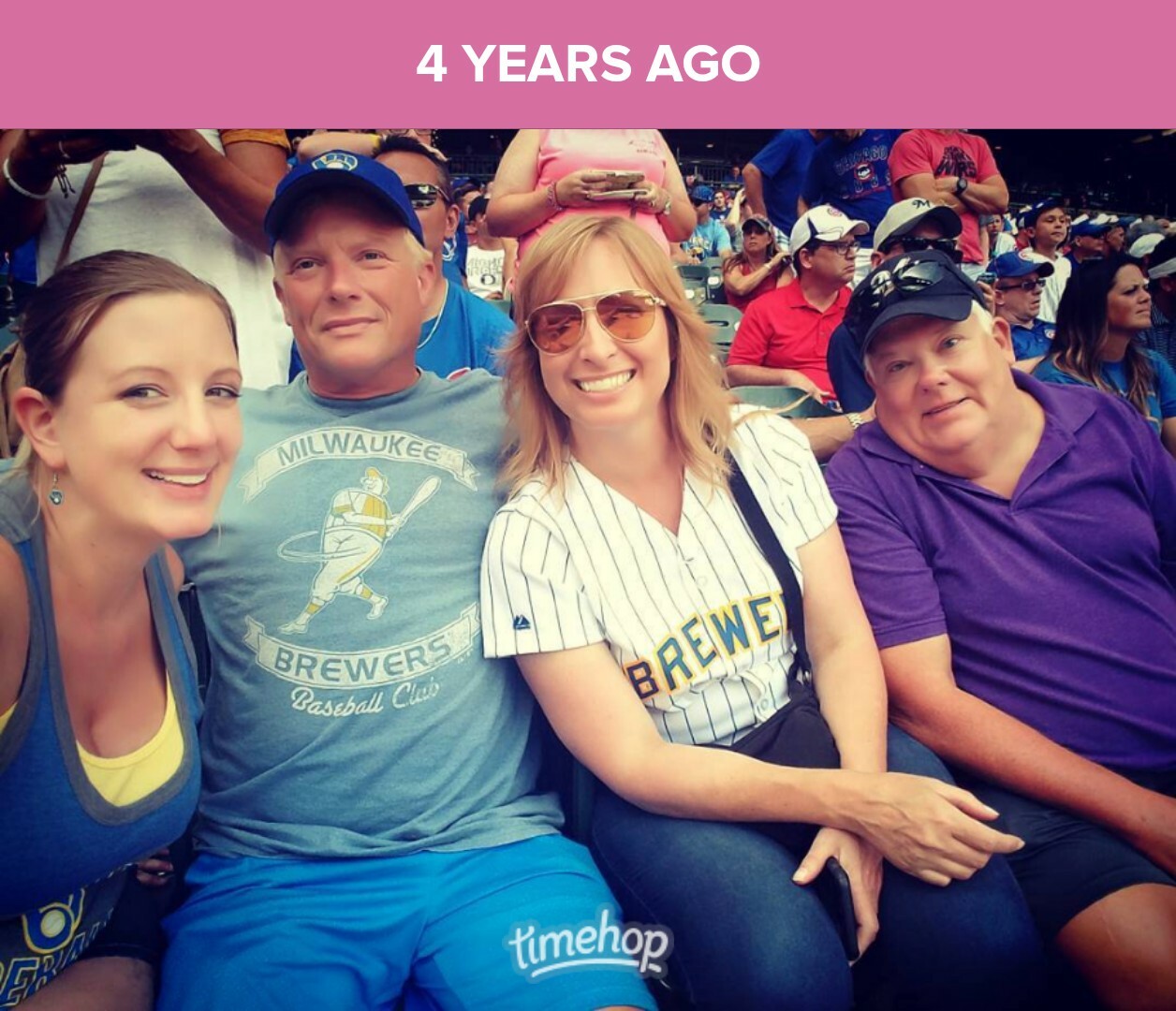 Four friends seated in stadium seats at a baseball game, smiling at the camera; two wearing Milwaukee Brewers shirts.