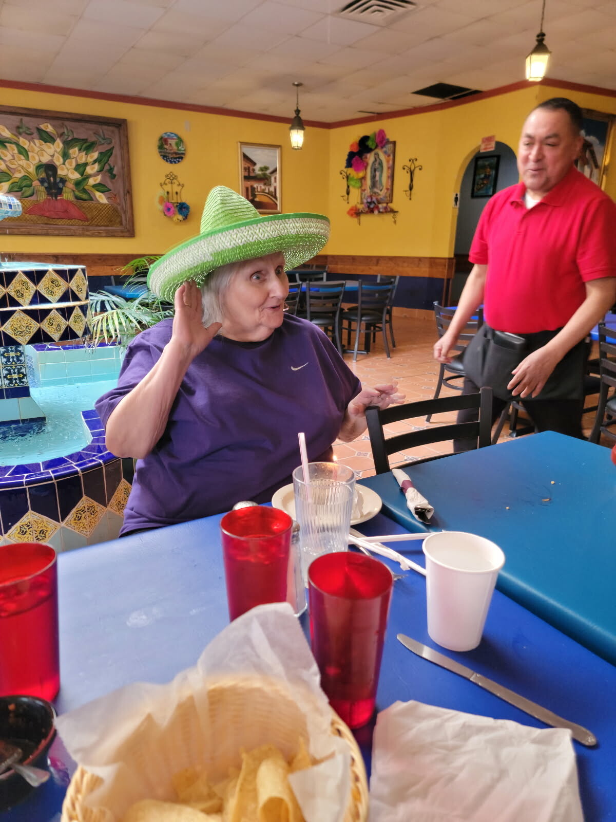 Older woman in a purple shirt wearing a green sombrero sits at a bright blue table in a colorful Mexican restaurant; a server stands nearby.