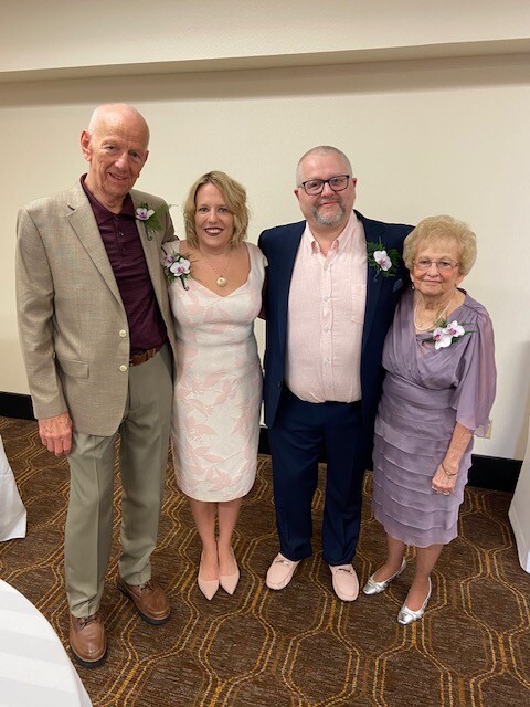 Four adults pose together at a formal event indoors, two men and two women wearing corsages, smiling at the camera.