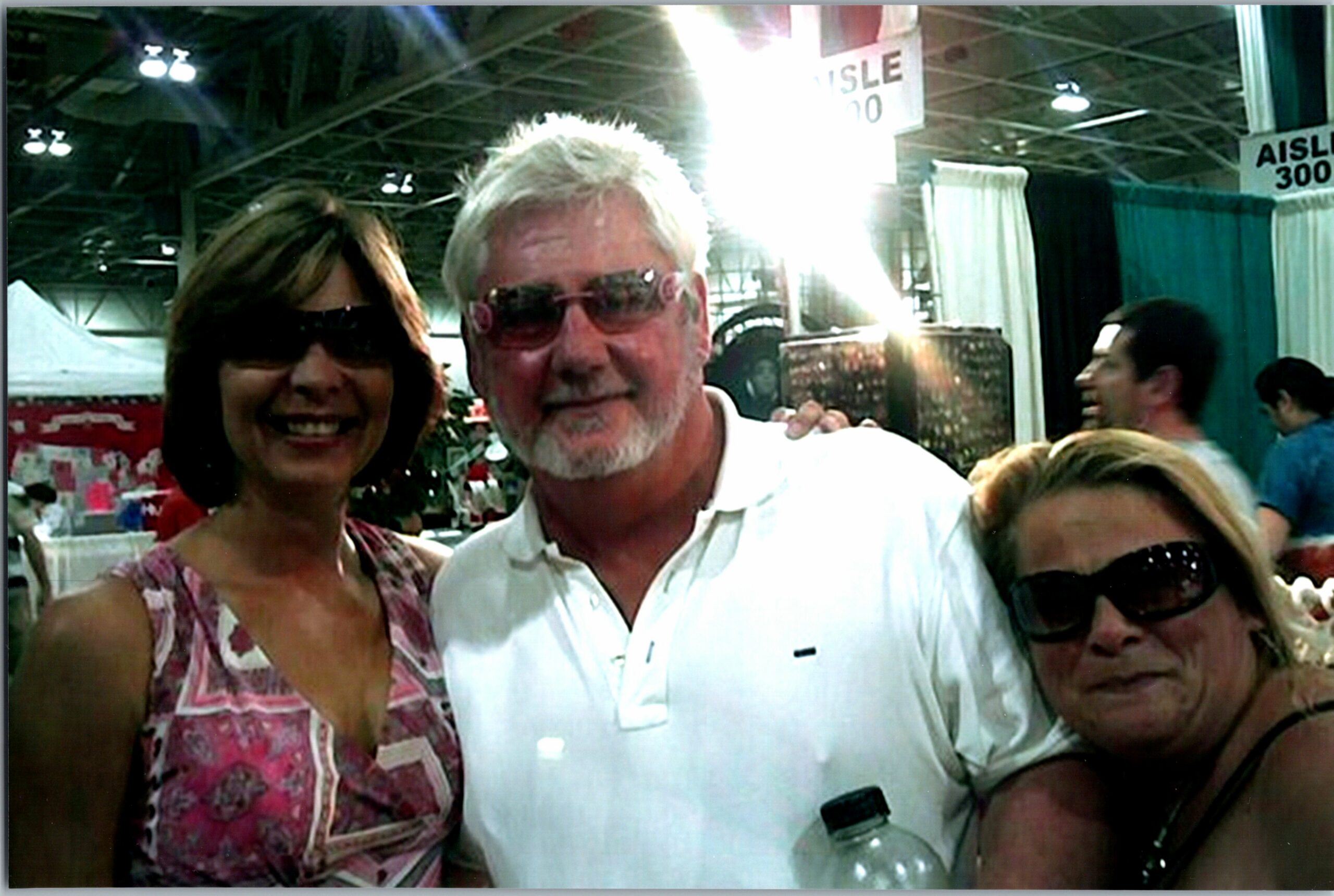 Three adults smiling for a photo at an indoor event; man with white hair and sunglasses center, woman in pink patterned top on left, woman with large sunglasses on right holding a water bottle.