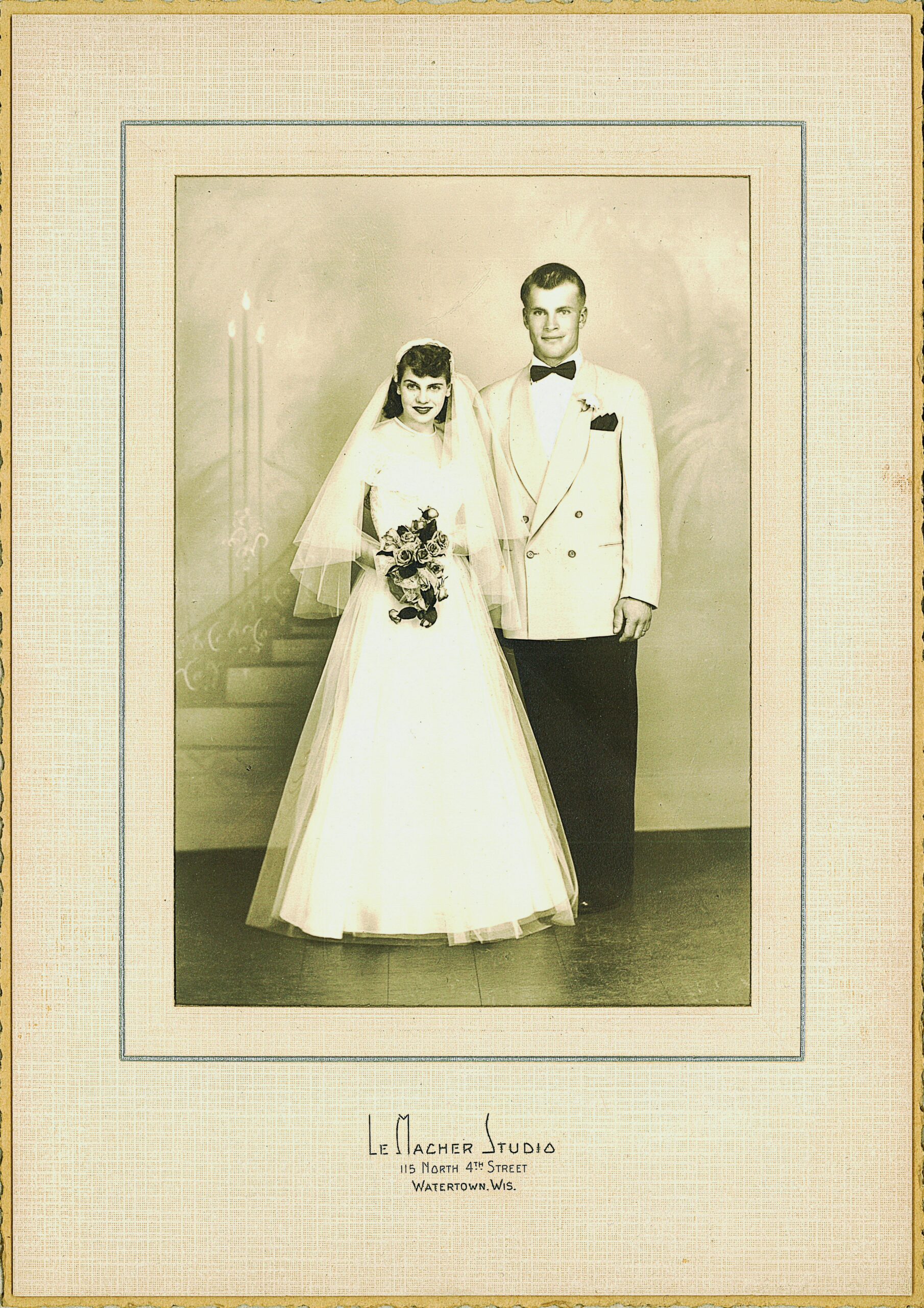 Bride in a white wedding dress and veil holding a bouquet, standing beside groom in a light suit and bow tie in a formal studio portrait.