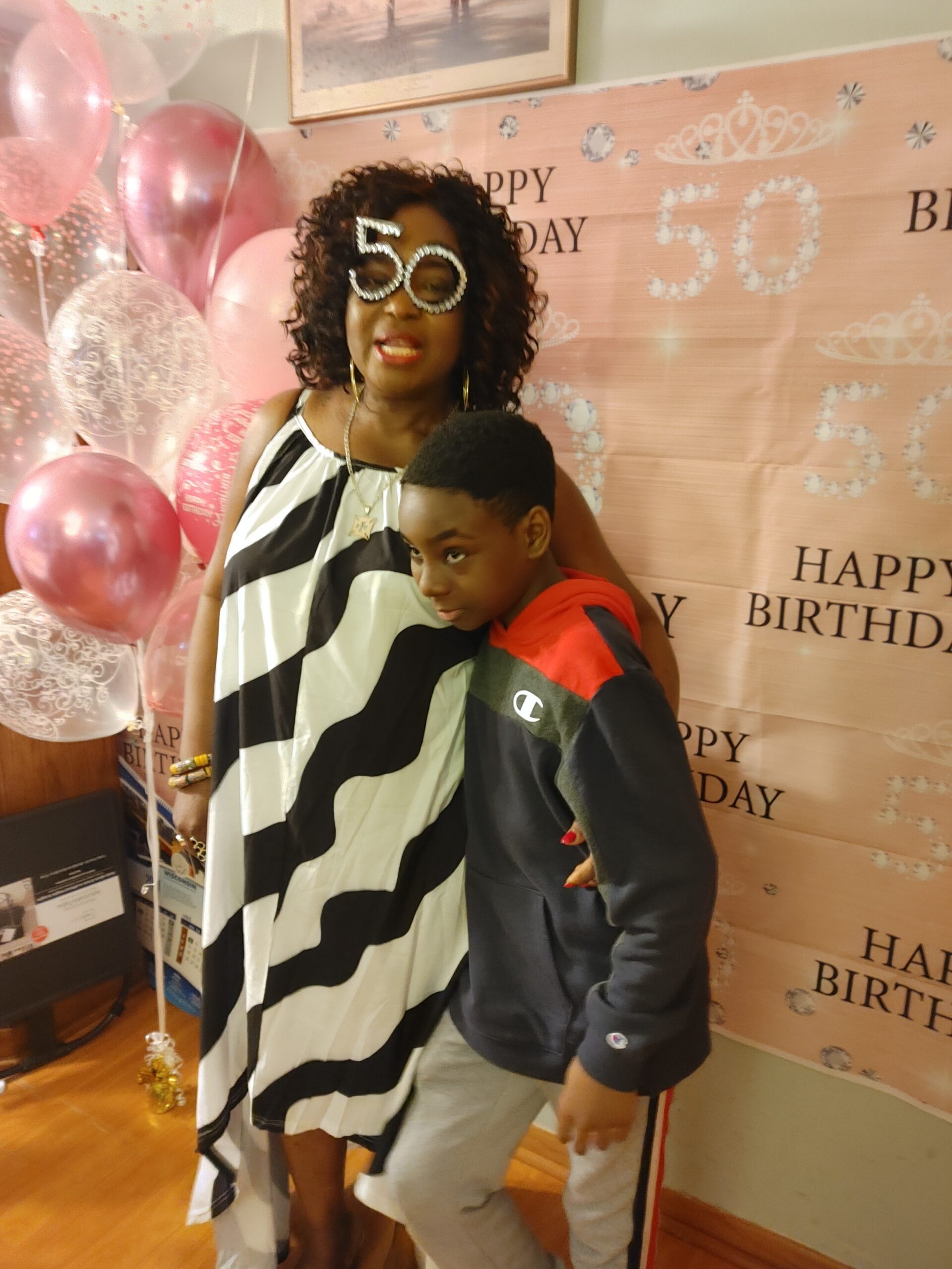 Woman wearing large '50' glasses and a black-and-white dress posing with a smiling boy at a birthday backdrop with balloons.
