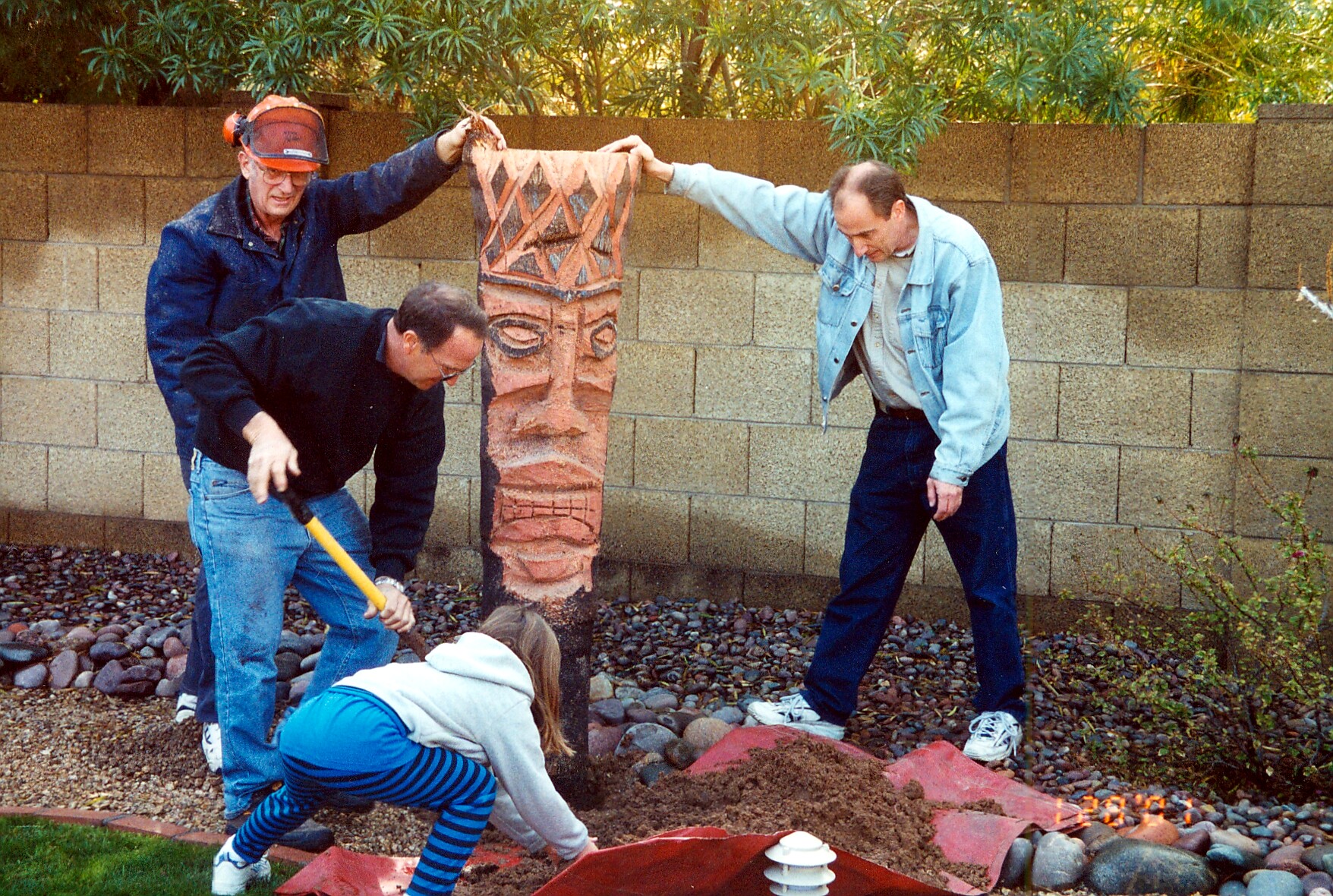 Group of adults and a child digging in a backyard garden around a carved totem-like statue.