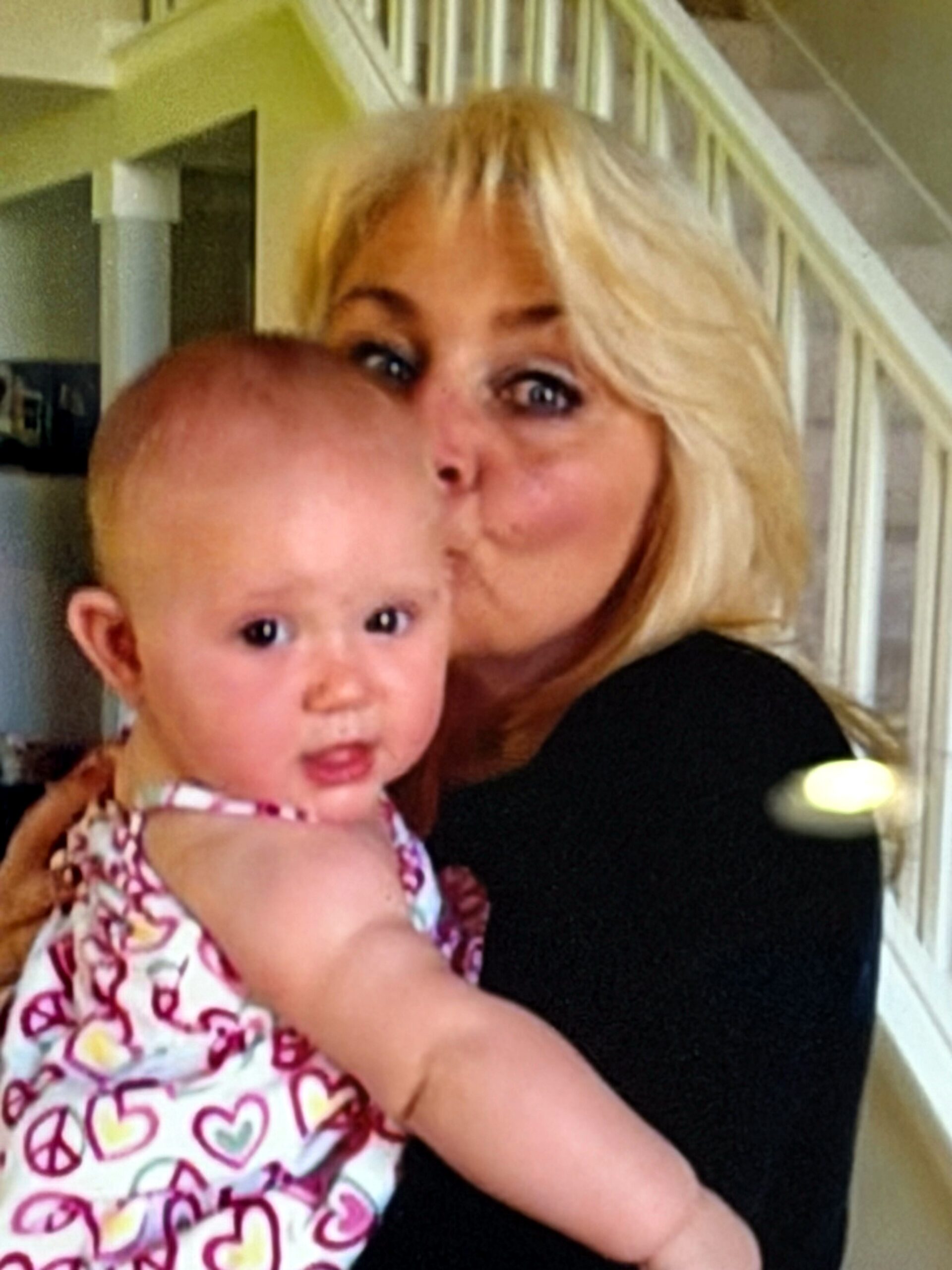 Woman with blonde hair smiling while holding a baby in a patterned dress indoors near a staircase.