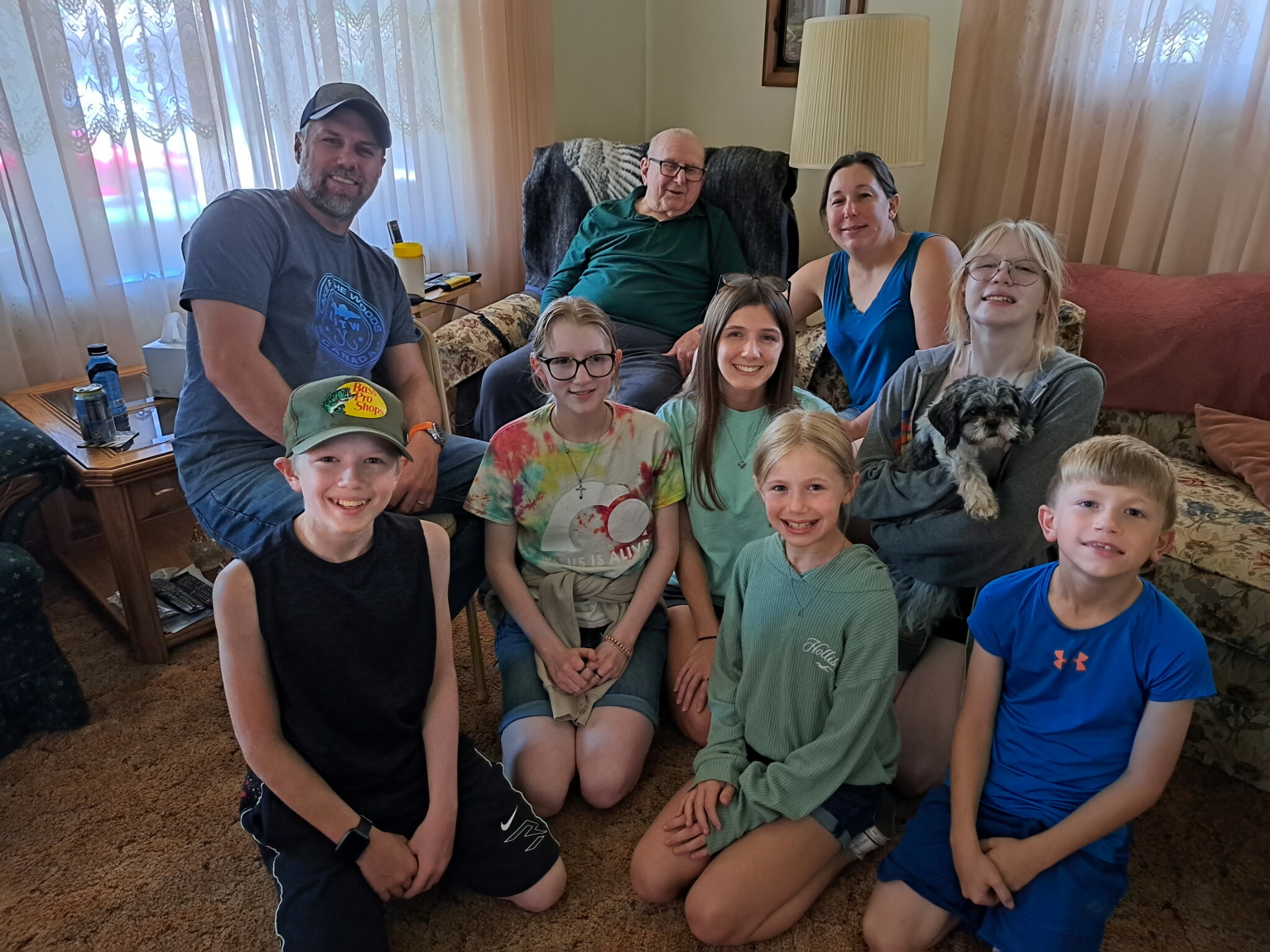 Smiling family group in a cozy living room: children seated on the floor and couch, adults behind them, and a small dog on a girl's lap; warm, homey setting with beige carpet and sheer curtains.