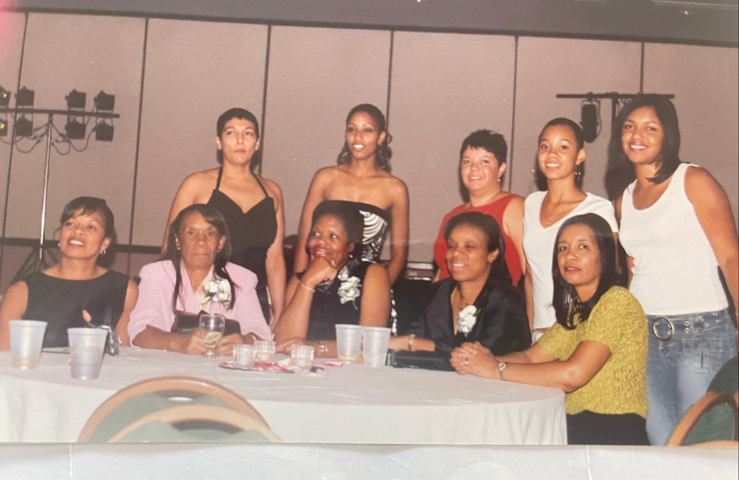 Group of women in dresses gathered around a table at a celebration, posing for a photo with drinks on the table and smiles all around.